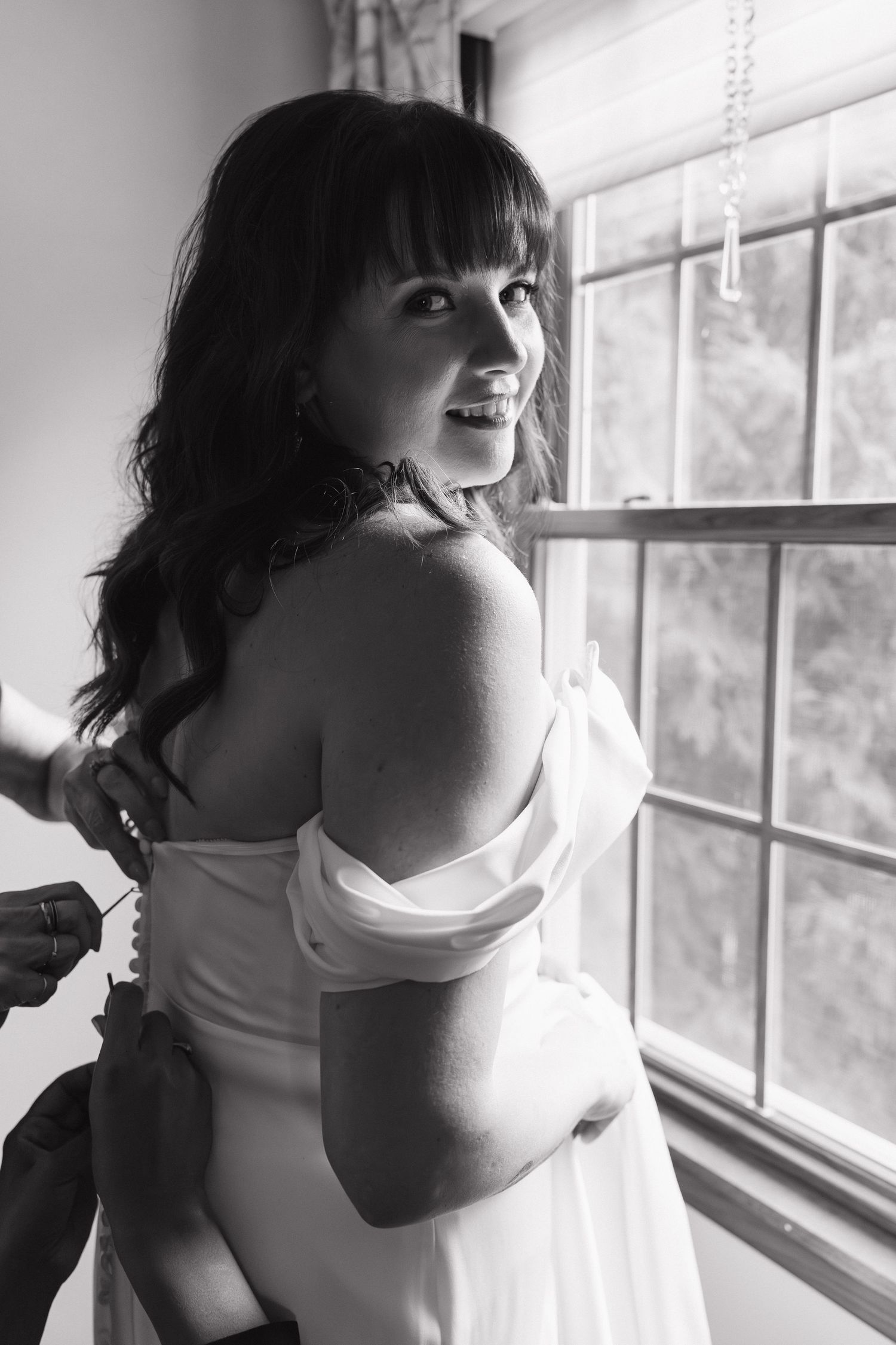 Black and white photo of smiling brunette bride in off-shoulder gown being buttoned up near window.