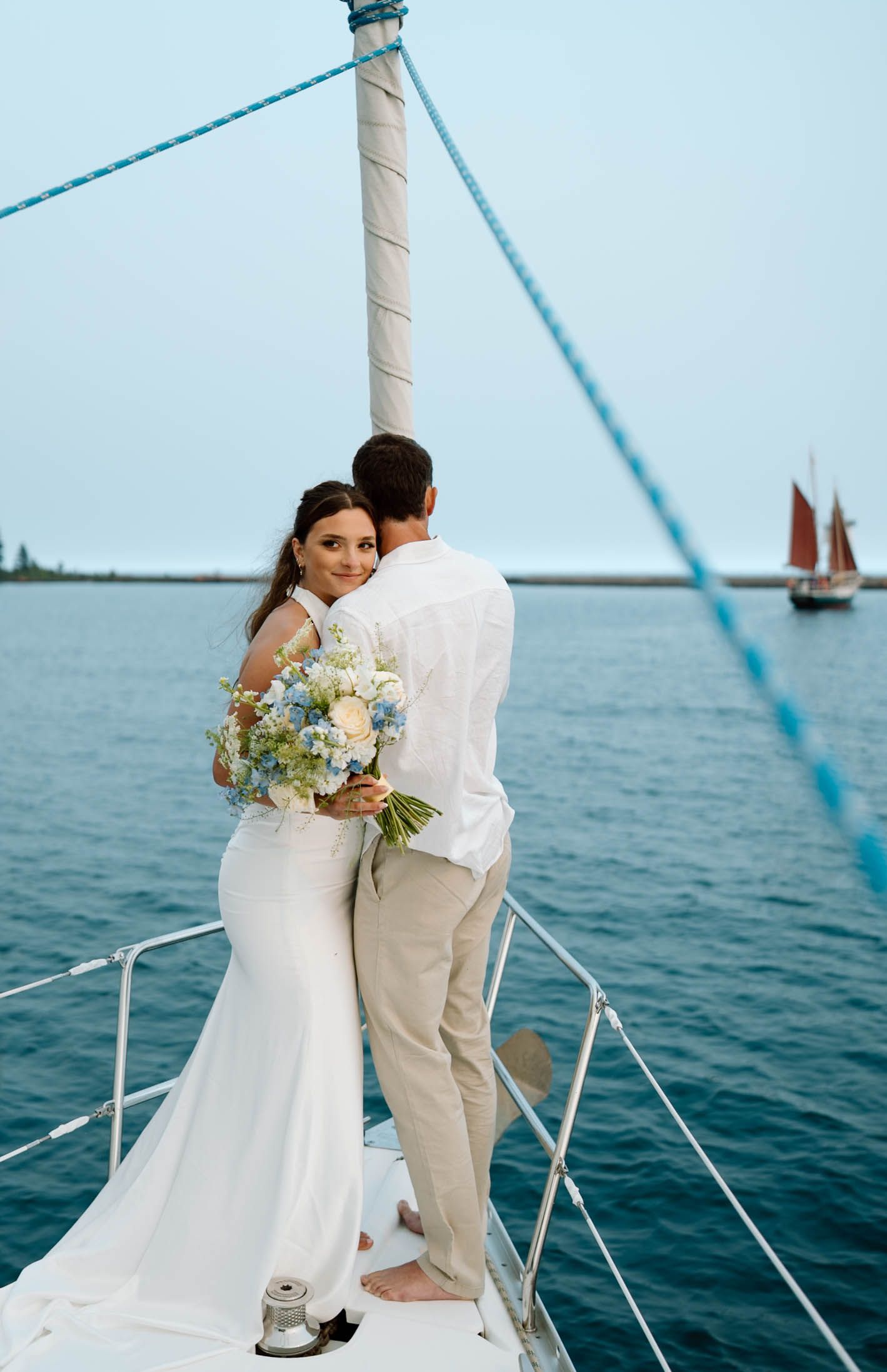 Bride and groom embracing on sailboat bow, bride holding blue and white bouquet, red sailboat visible in background.