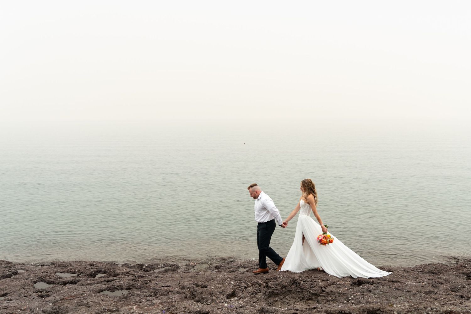 Bride and groom walk along rocky shoreline in misty coastal setting, bride holding orange bouquet in elegant white gown.