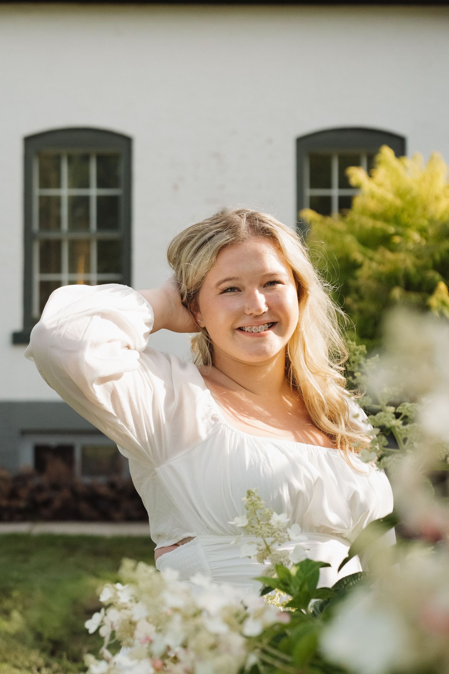 Smiling blonde woman in white top posing near white building with flowers.