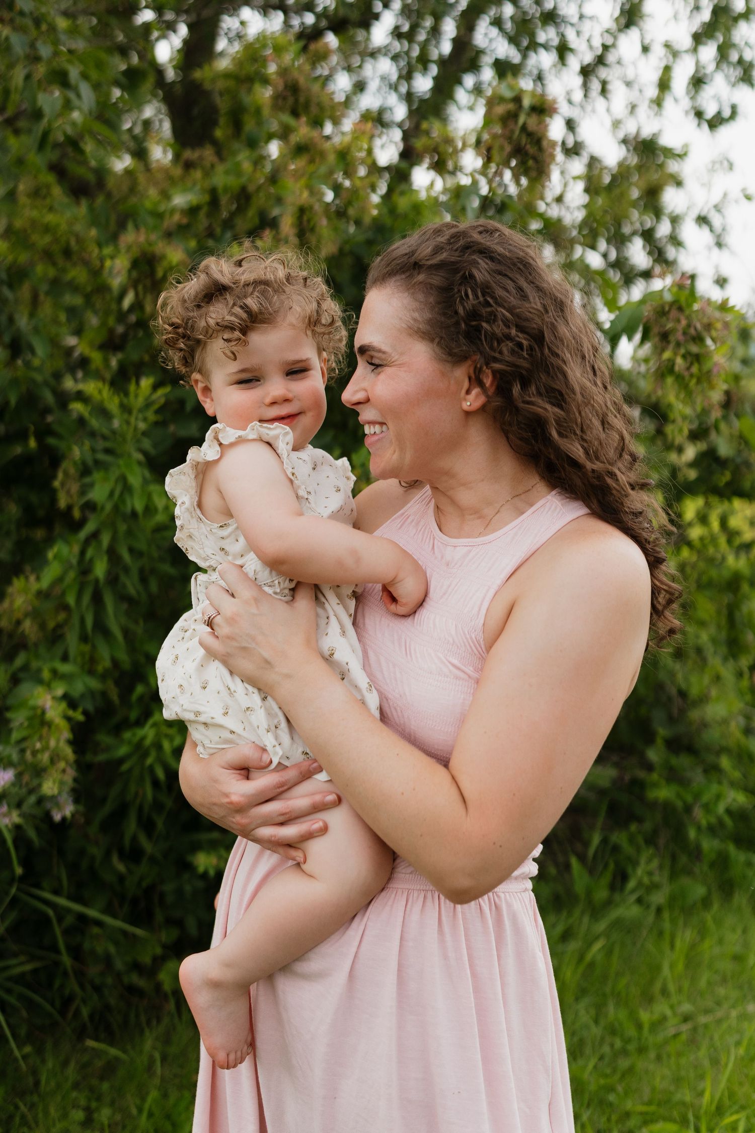 A smiling mother in a pink dress holds her curly-haired toddler in a white floral dress outdoors.