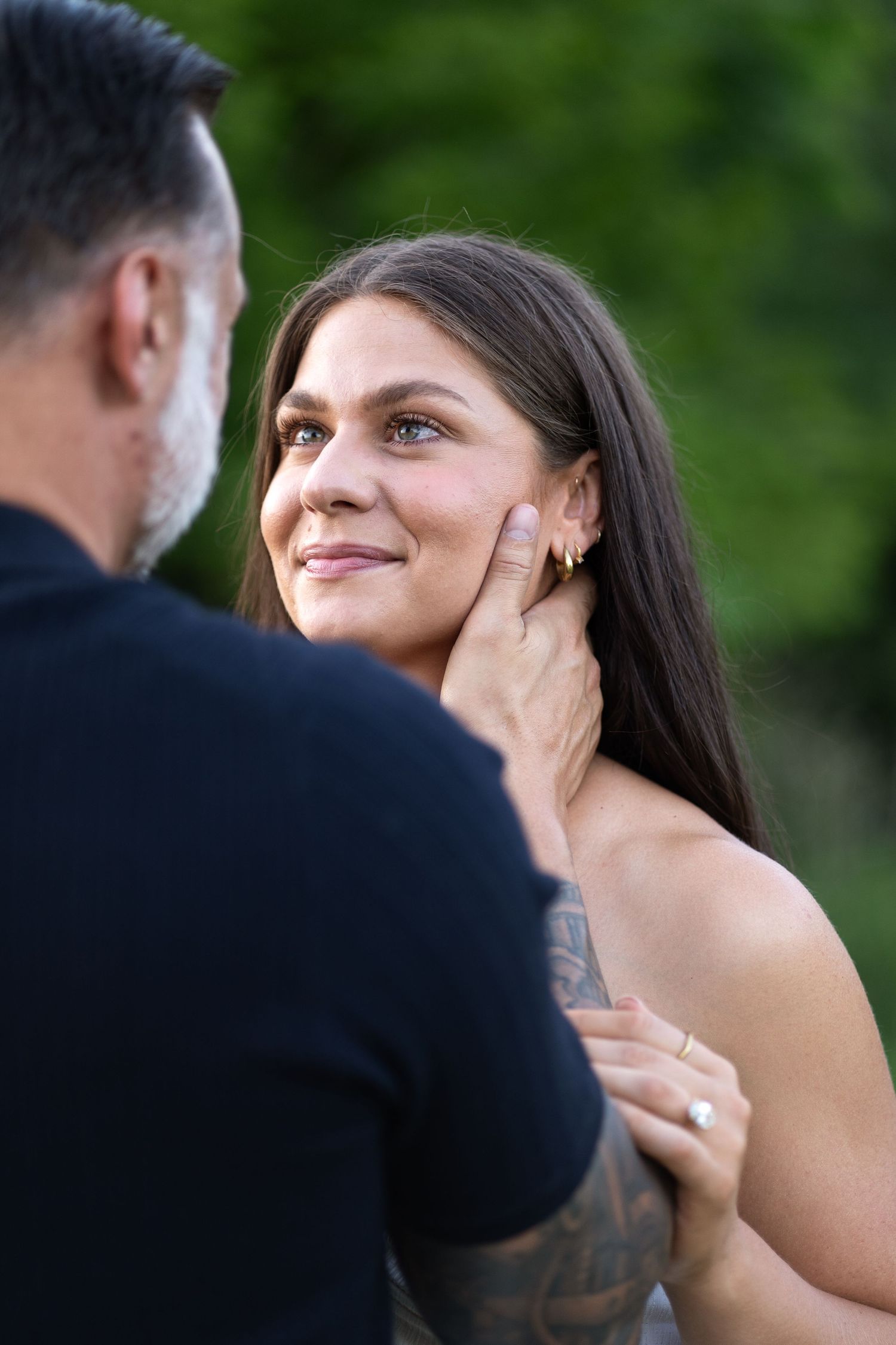 A tattooed man tenderly holds a smiling brunette woman's face outdoors with green foliage background.