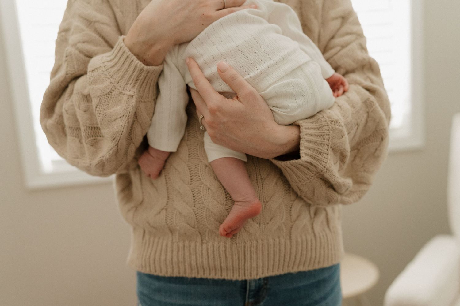 Parent in beige cable-knit sweater cradling a newborn baby against their chest in a bright, soft-lit room.