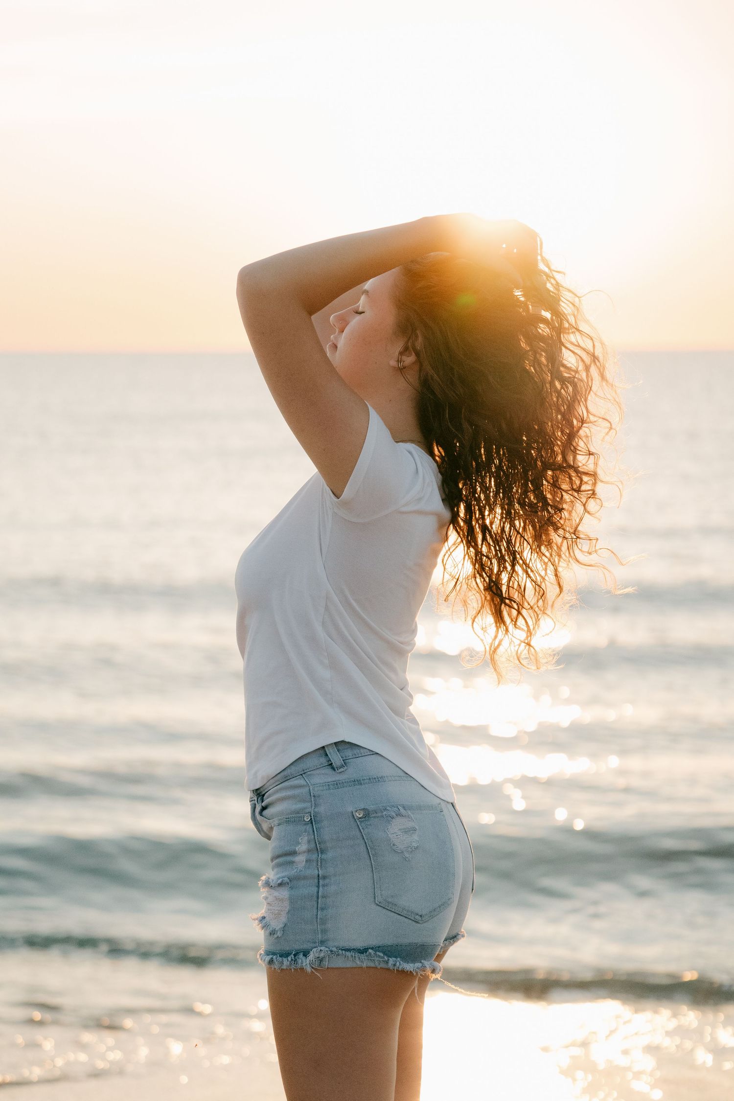 Young woman in white tee and denim shorts stands at beach during stunning sunset.