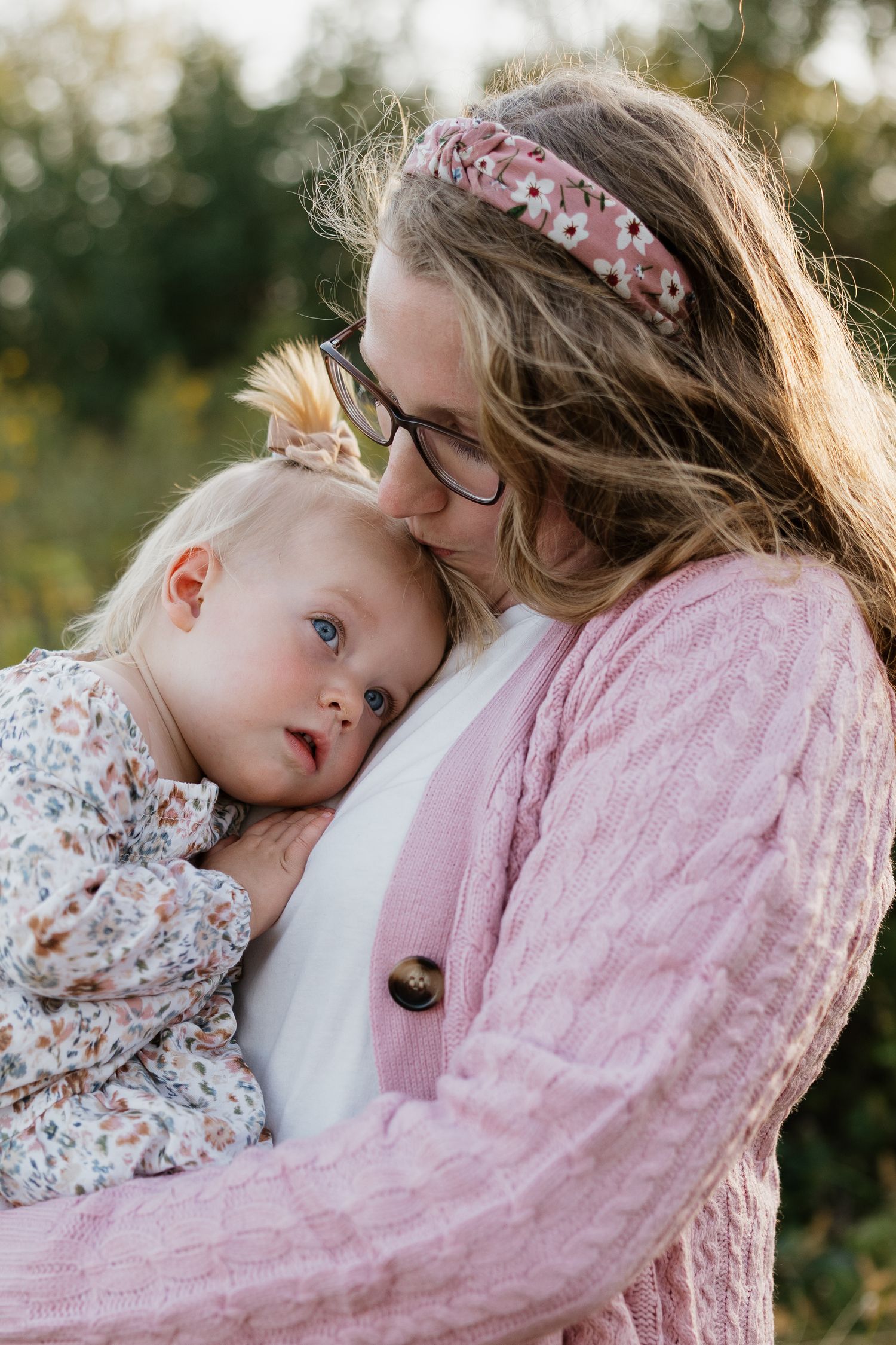 Tender moment of a mother in pink cardigan kissing her baby girl outdoors in a natural setting.