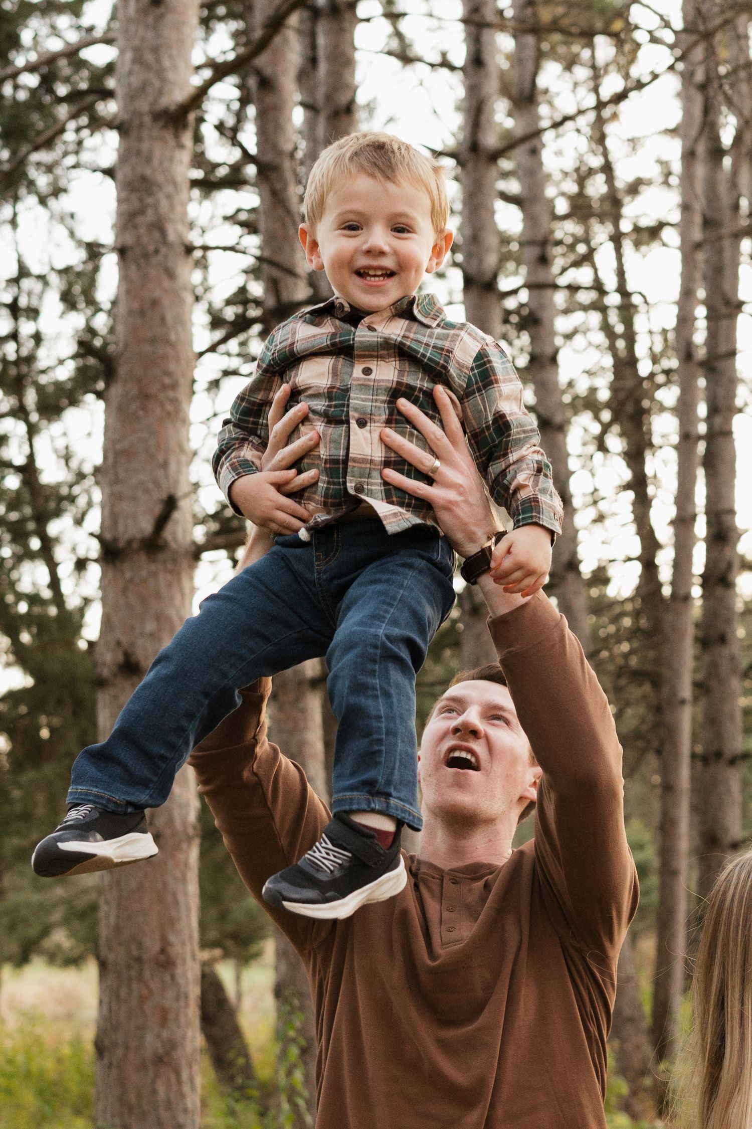 Excited father lifting laughing toddler boy high in the air among tall pine trees outdoors.