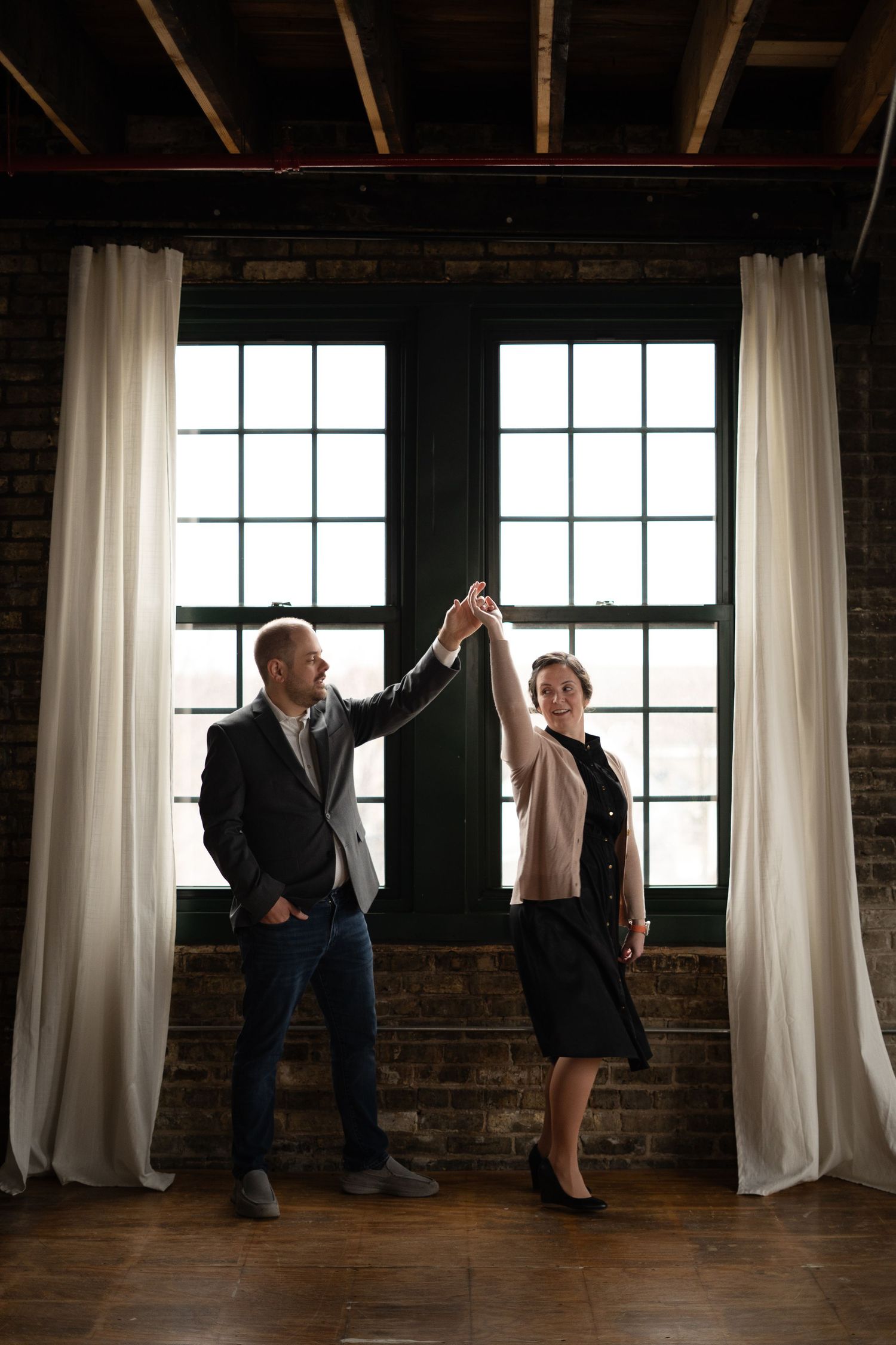 Couple dancing together in front of large windows with white curtains in loft space.