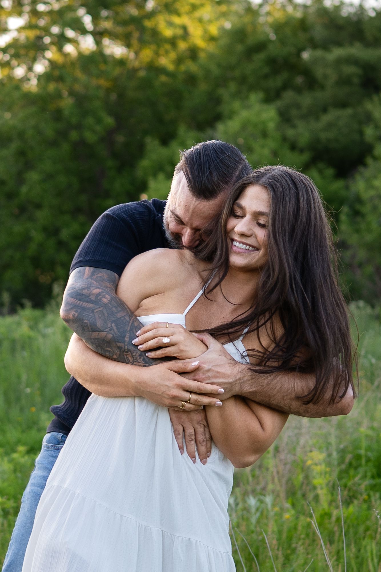 A couple embraces outdoors, woman in white dress laughing joyfully as tattooed man hugs her from behind in a green field.