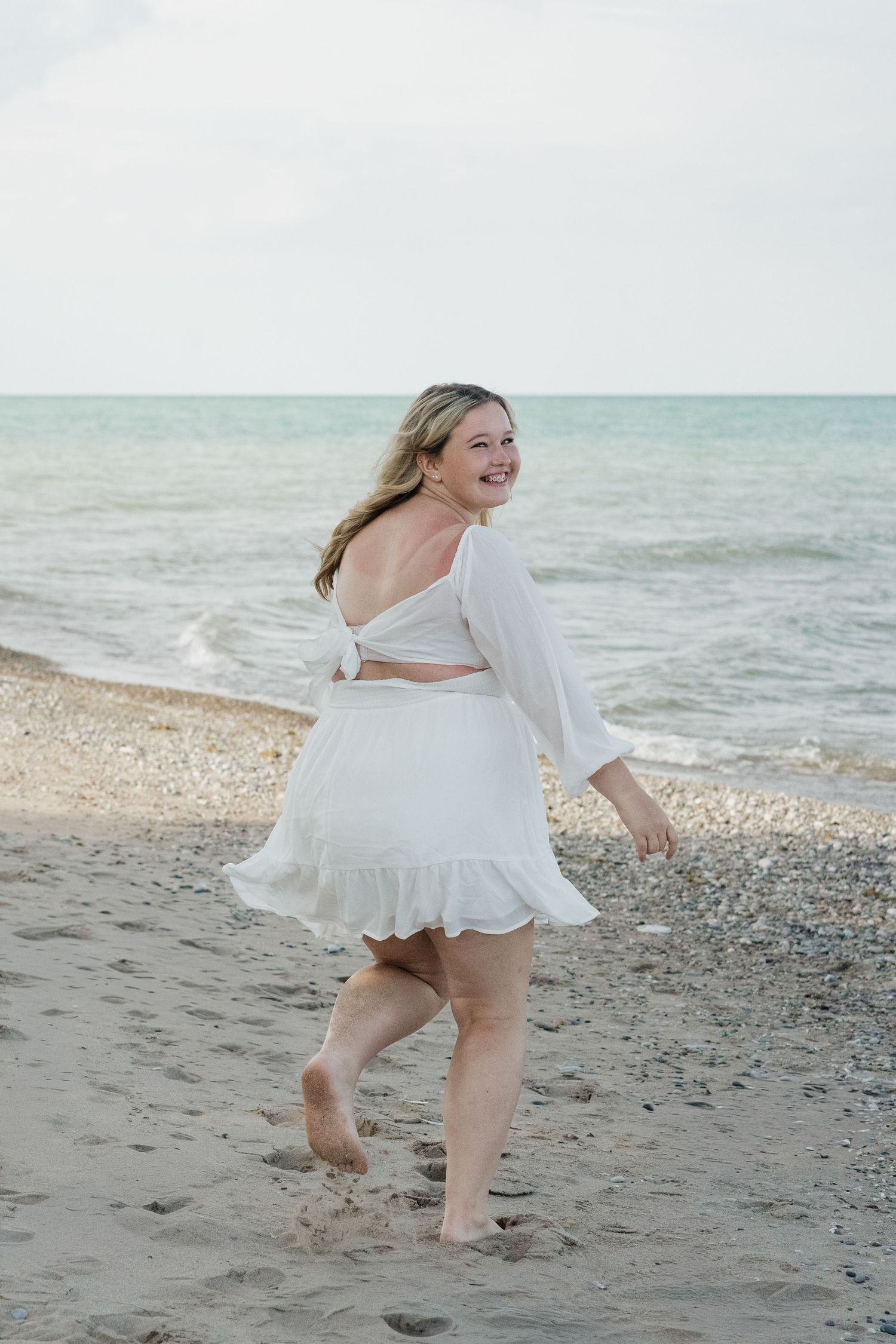 Blonde woman in white crop top and skirt walking barefoot on sandy beach.