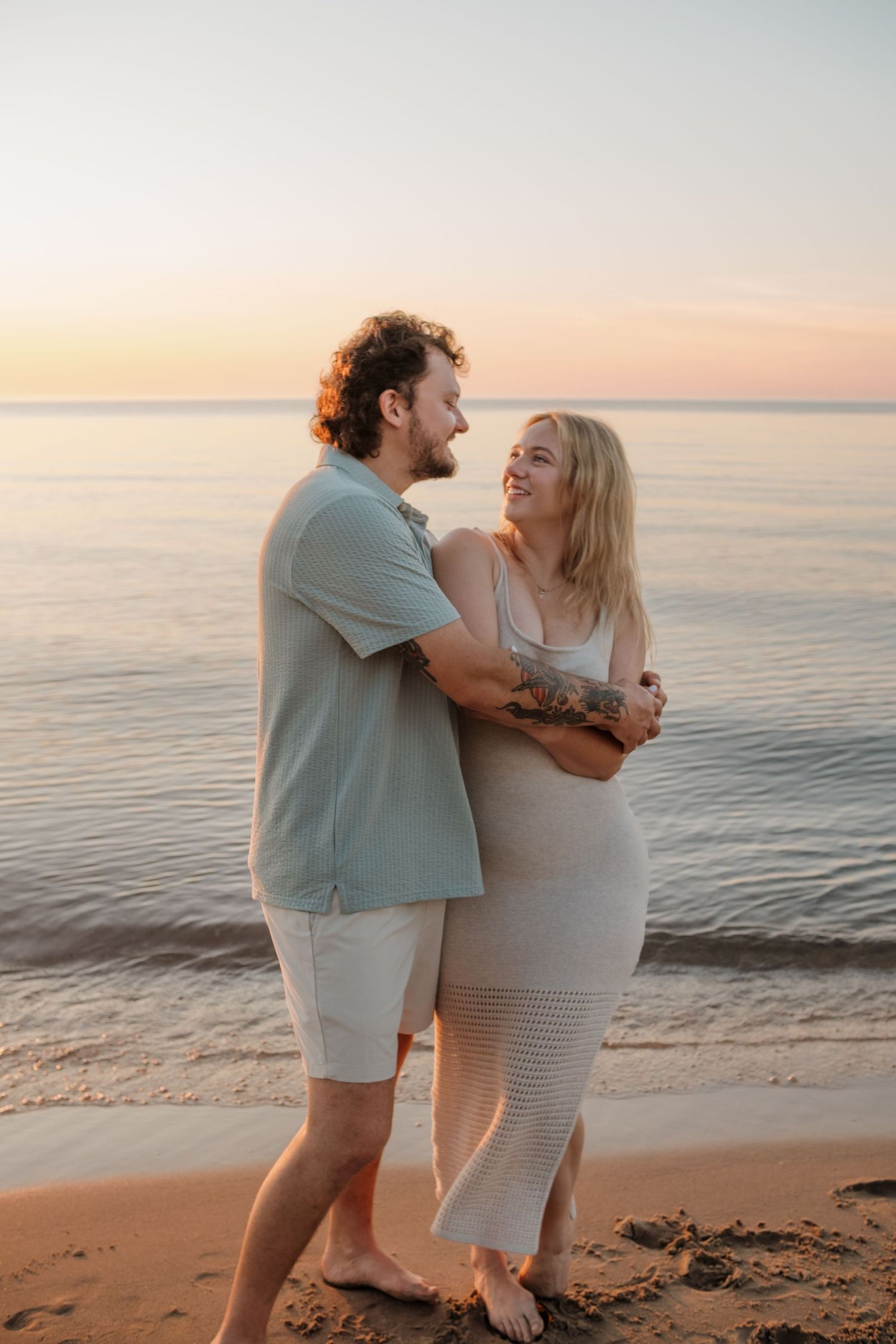 Happy couple embracing on sandy beach at sunset, woman in cream knit dress and man in sage polo shirt.
