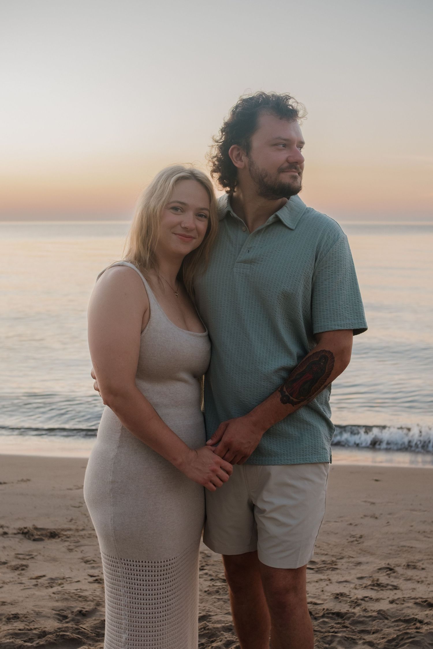 A couple holds hands and stands together on a sandy beach during a warm pastel sunset.