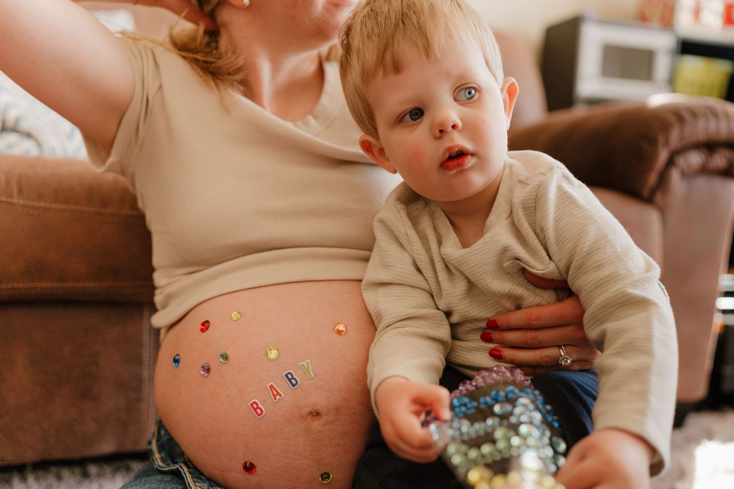 Pregnant woman sitting with toddler boy who holds toys, colorful stickers decorating her baby bump.