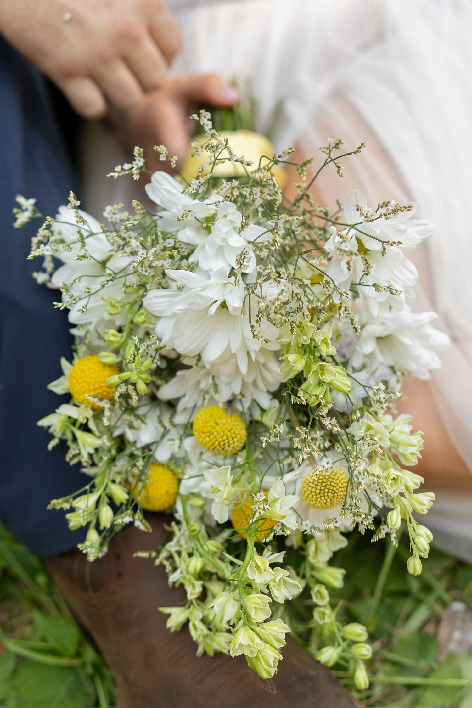 Rustic wedding bouquet with white daisies, yellow billy buttons, and wildflowers held by bride in white dress.