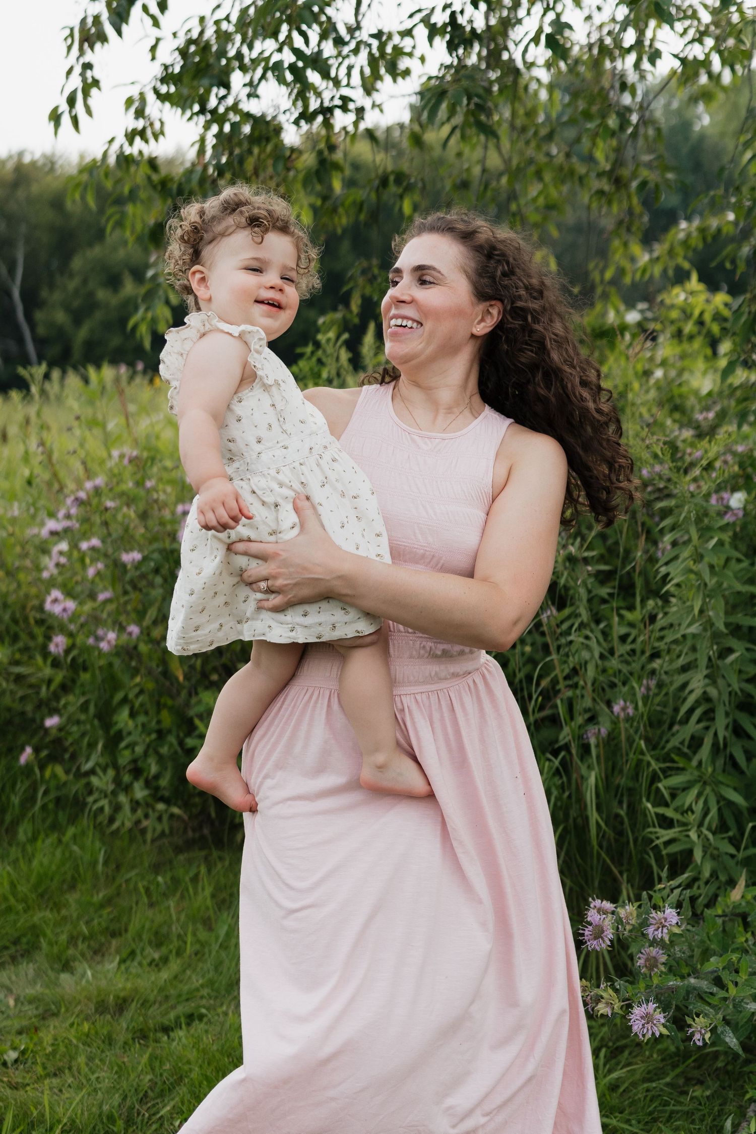 A joyful mother in a pink dress holds her laughing curly-haired toddler in a garden setting.
