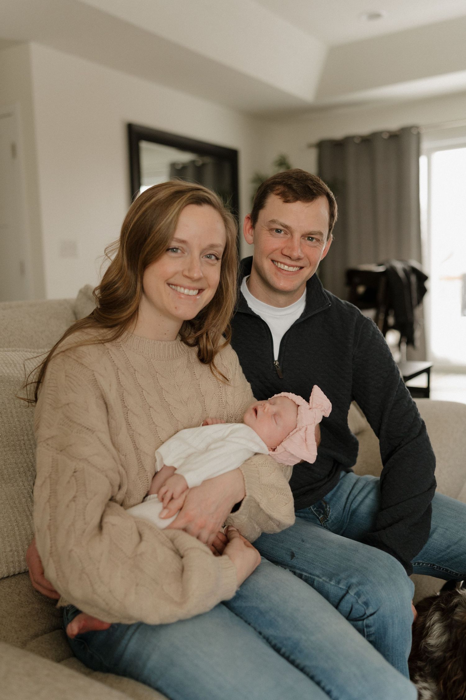 Smiling couple sitting on a couch holding their newborn baby dressed in pink at home.