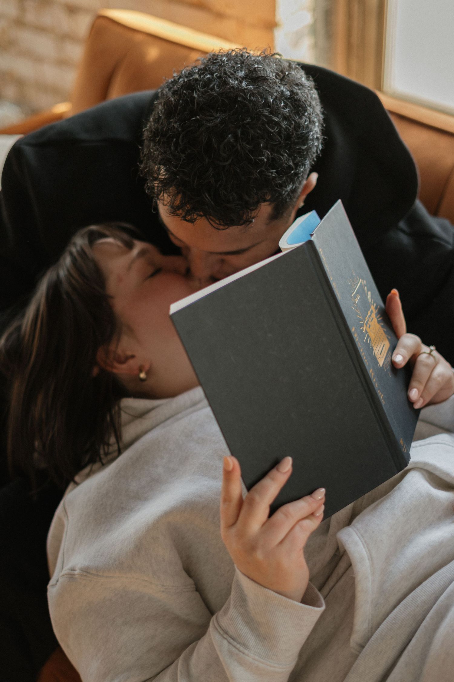 Romantic couple sharing a kiss behind a book while relaxing together on a couch indoors.