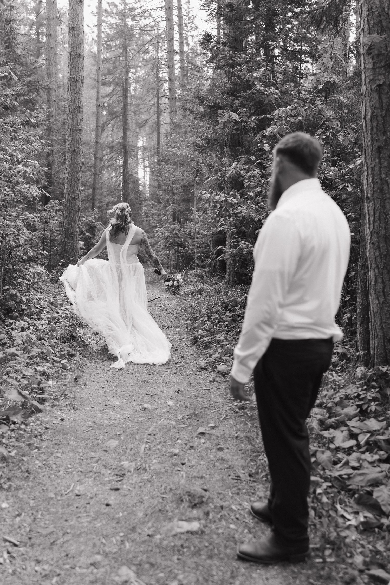 Black and white photo of bride in flowing gown walking forest path as groom watches during wedding day.
