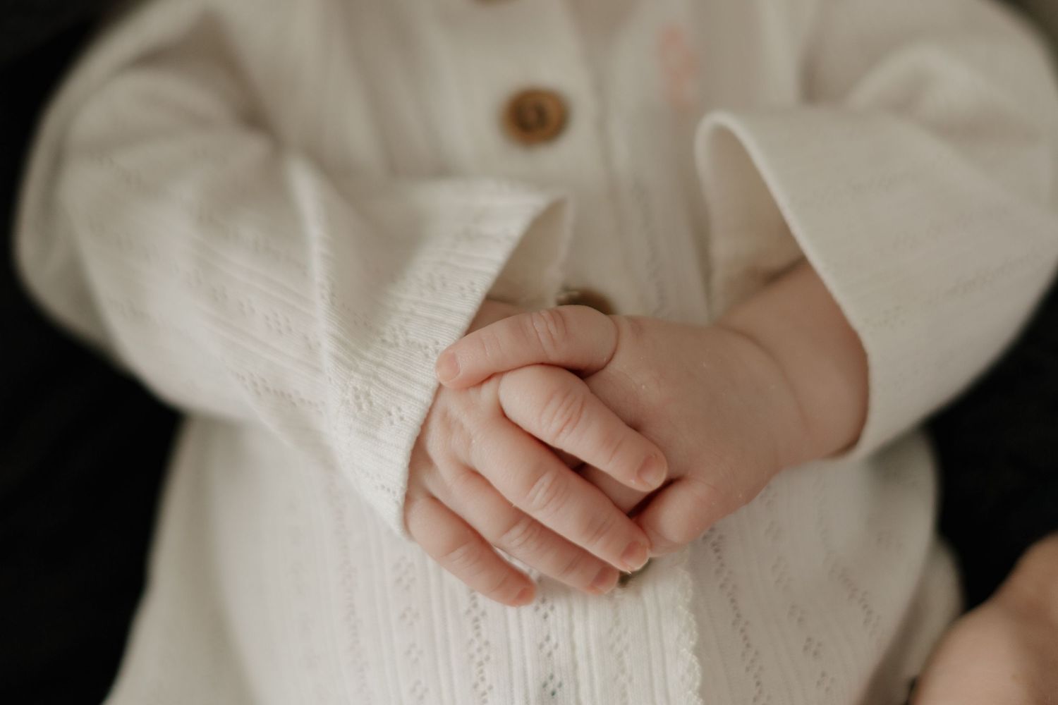 Close-up of a baby's clasped hands resting on a white knit outfit with button detail.
