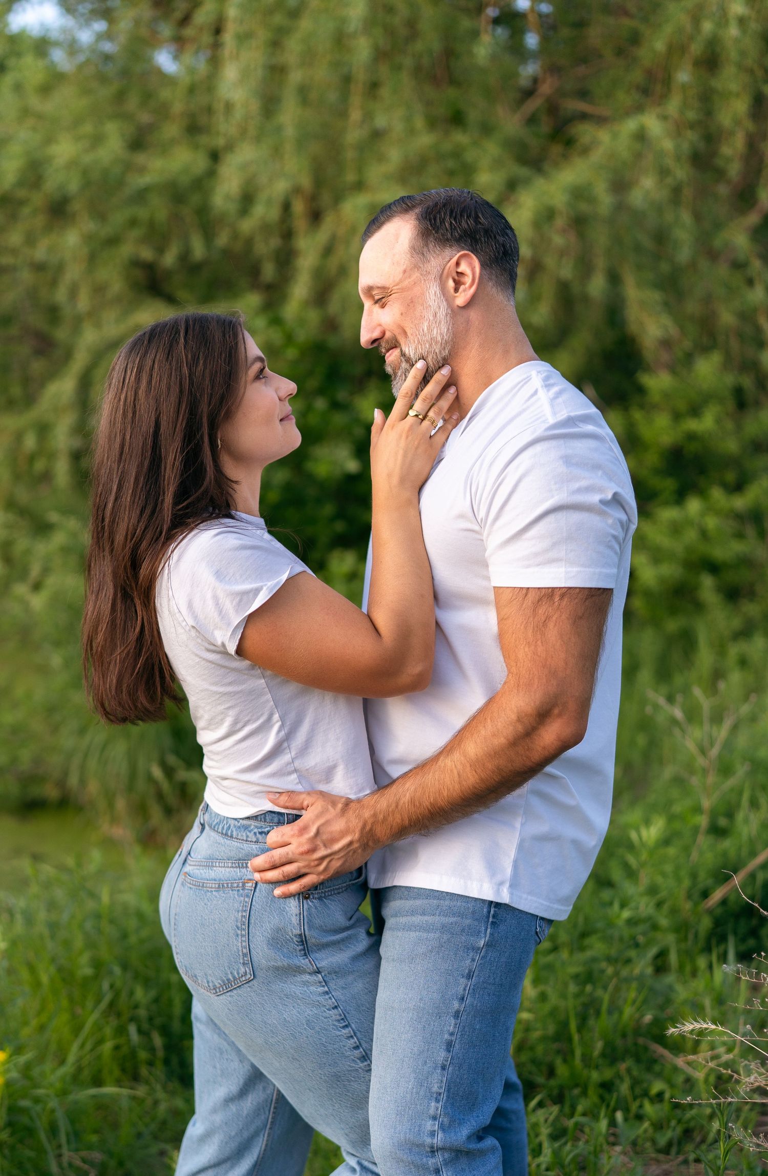 A couple in matching white t-shirts and jeans embrace outdoors, gazing at each other with green trees behind them.