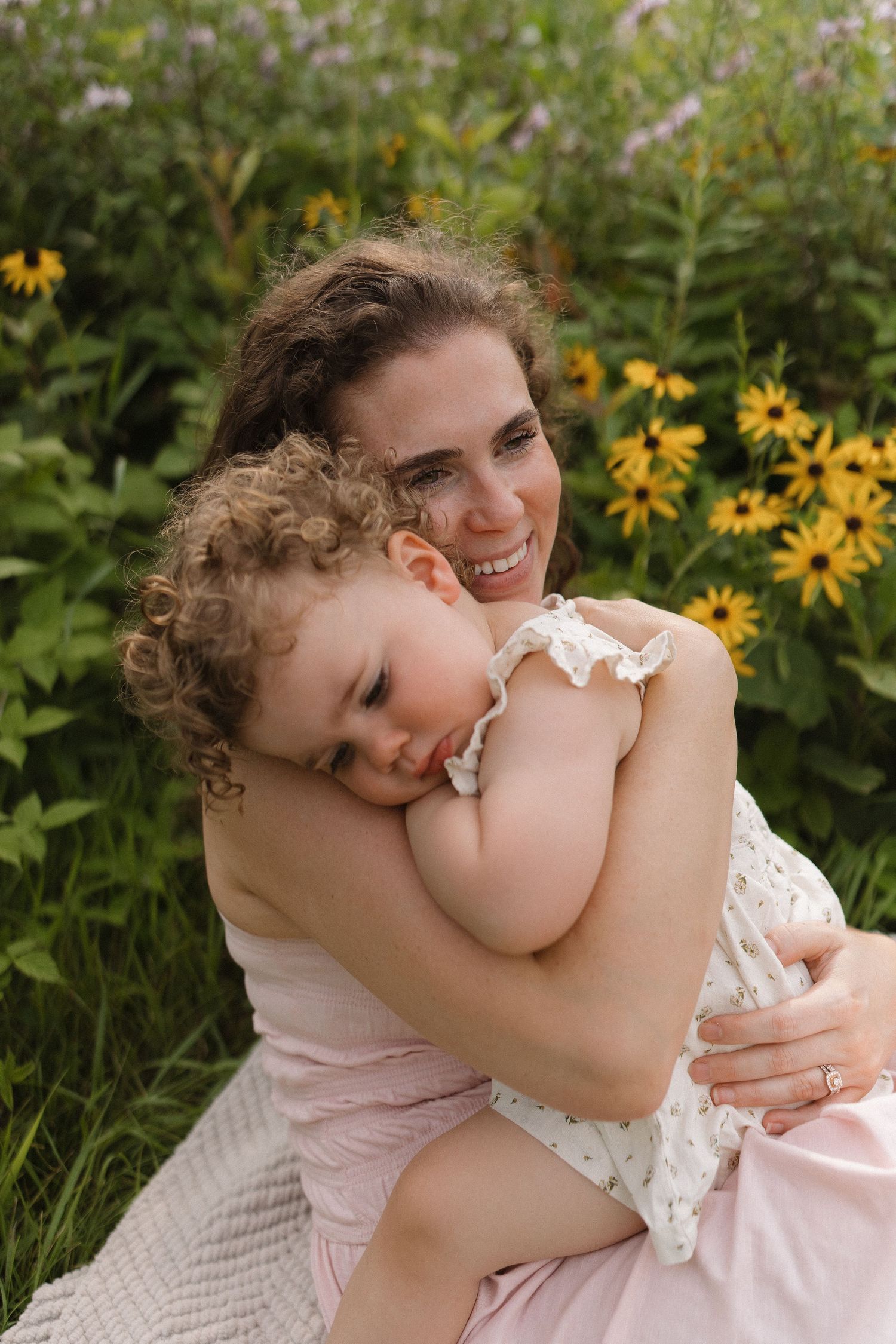 Smiling mother embraces curly-haired toddler daughter on blanket surrounded by yellow wildflowers.