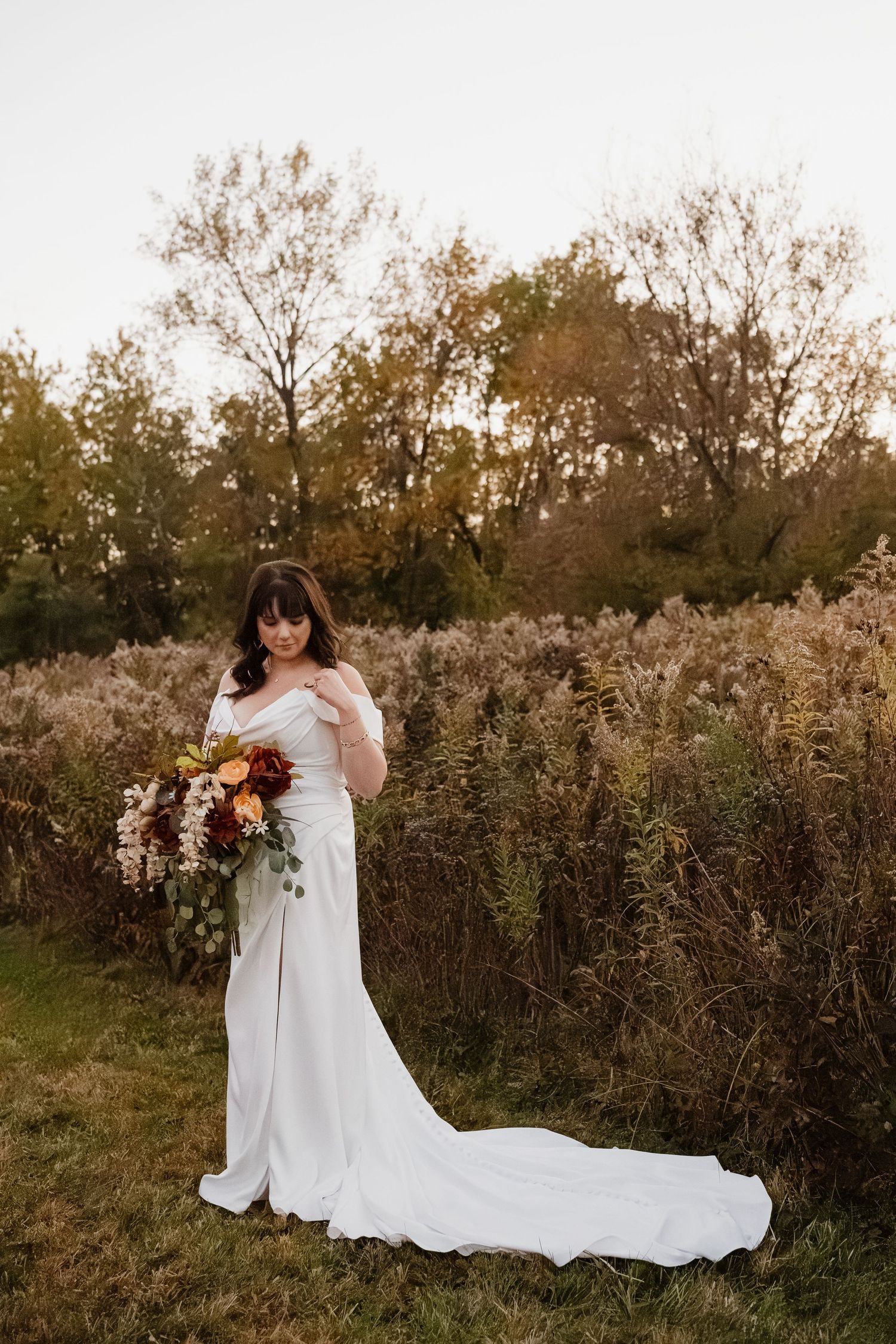 Bride in off-shoulder white gown with train holds autumn bouquet in golden fall outdoor landscape.