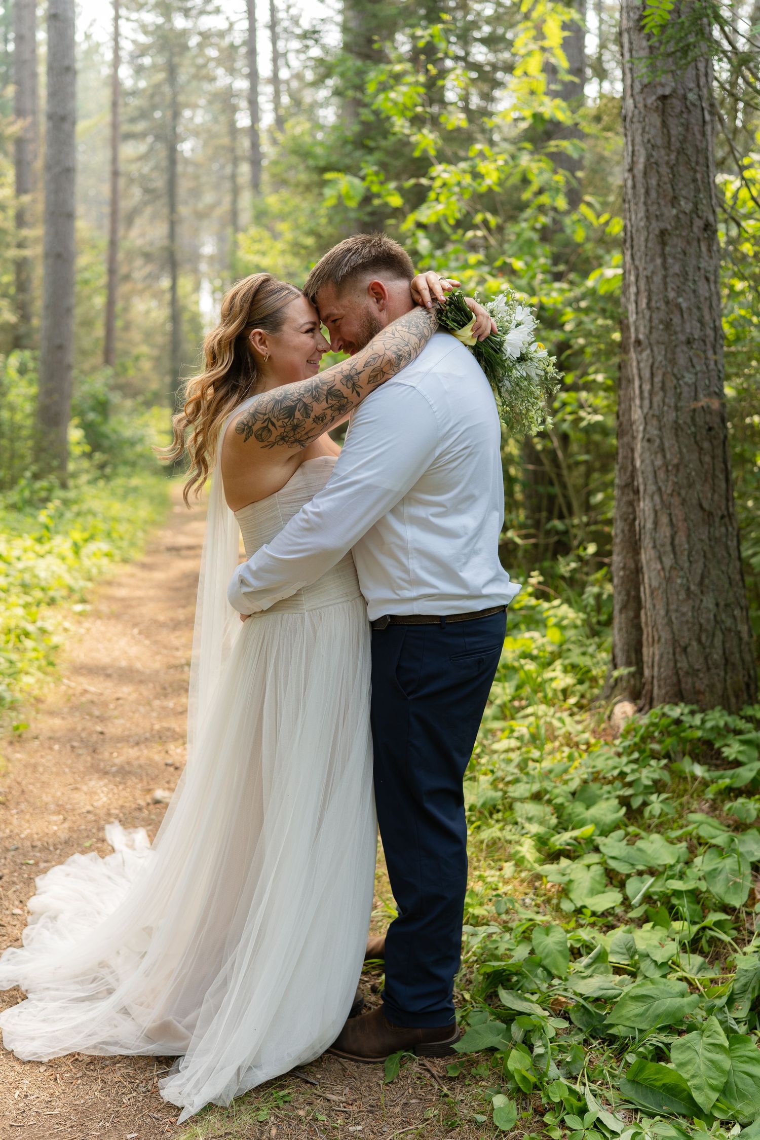 A bride and groom share a romantic embrace on a forest path, the bride in a flowing white gown holding a bouquet.