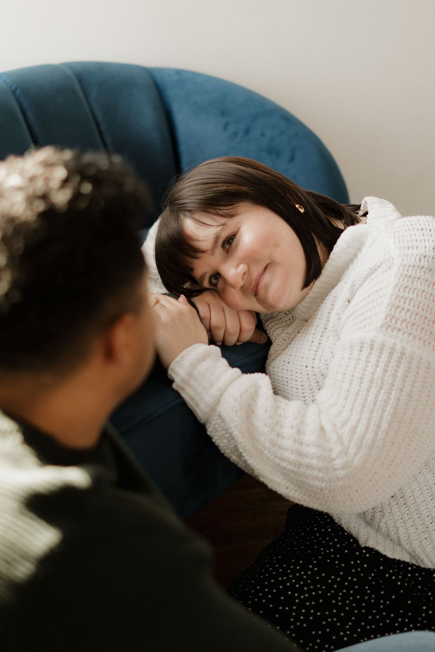 A woman in a white sweater rests her head on a blue chair, holding hands with a person sitting nearby.