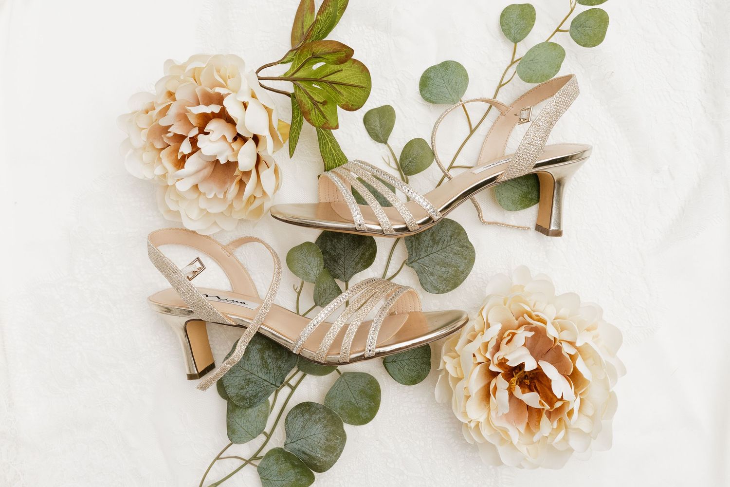 Elegant beige strappy heeled sandals displayed with peach roses and eucalyptus on white background.