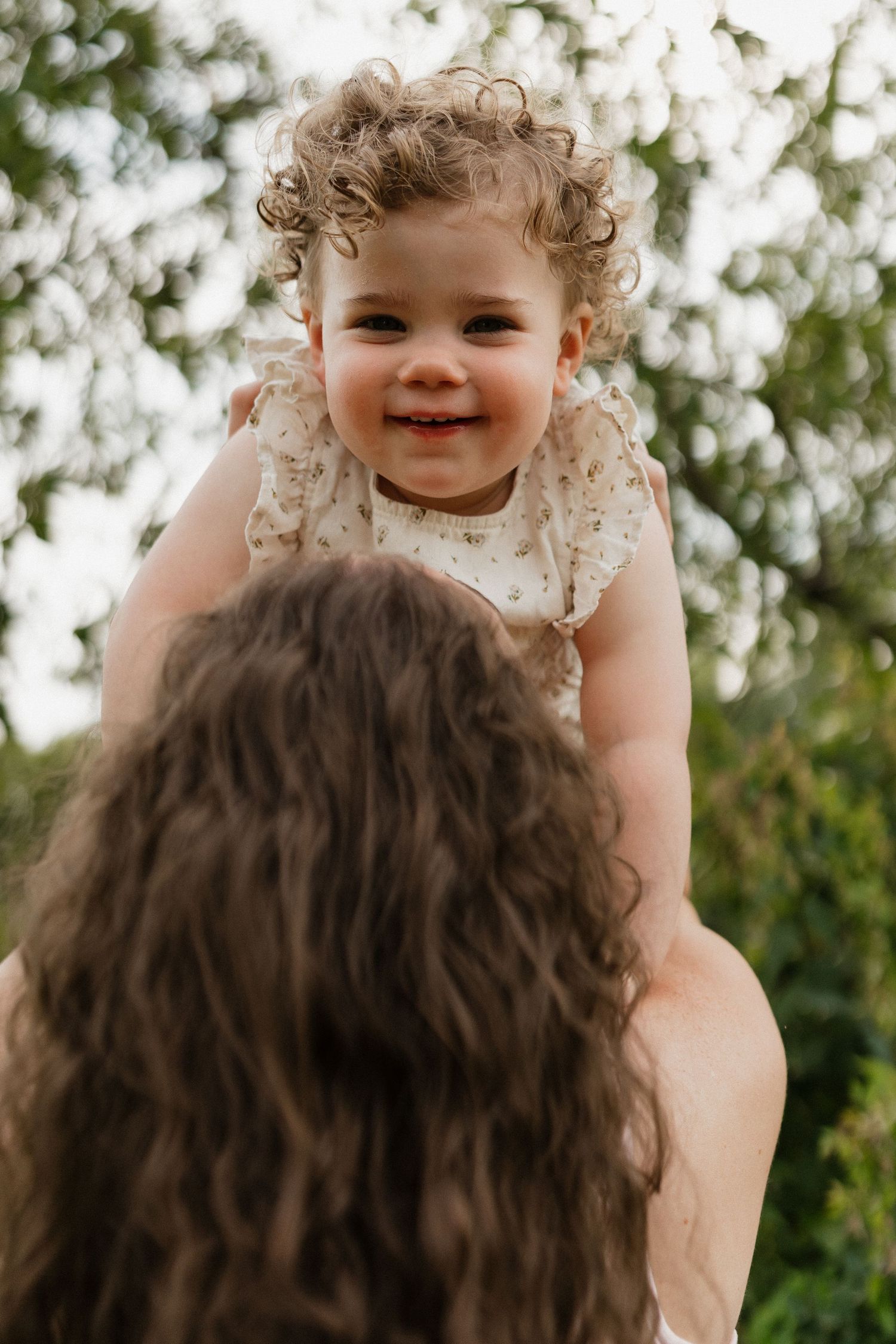 Laughing curly-haired baby girl lifted up by mother with green trees in background.