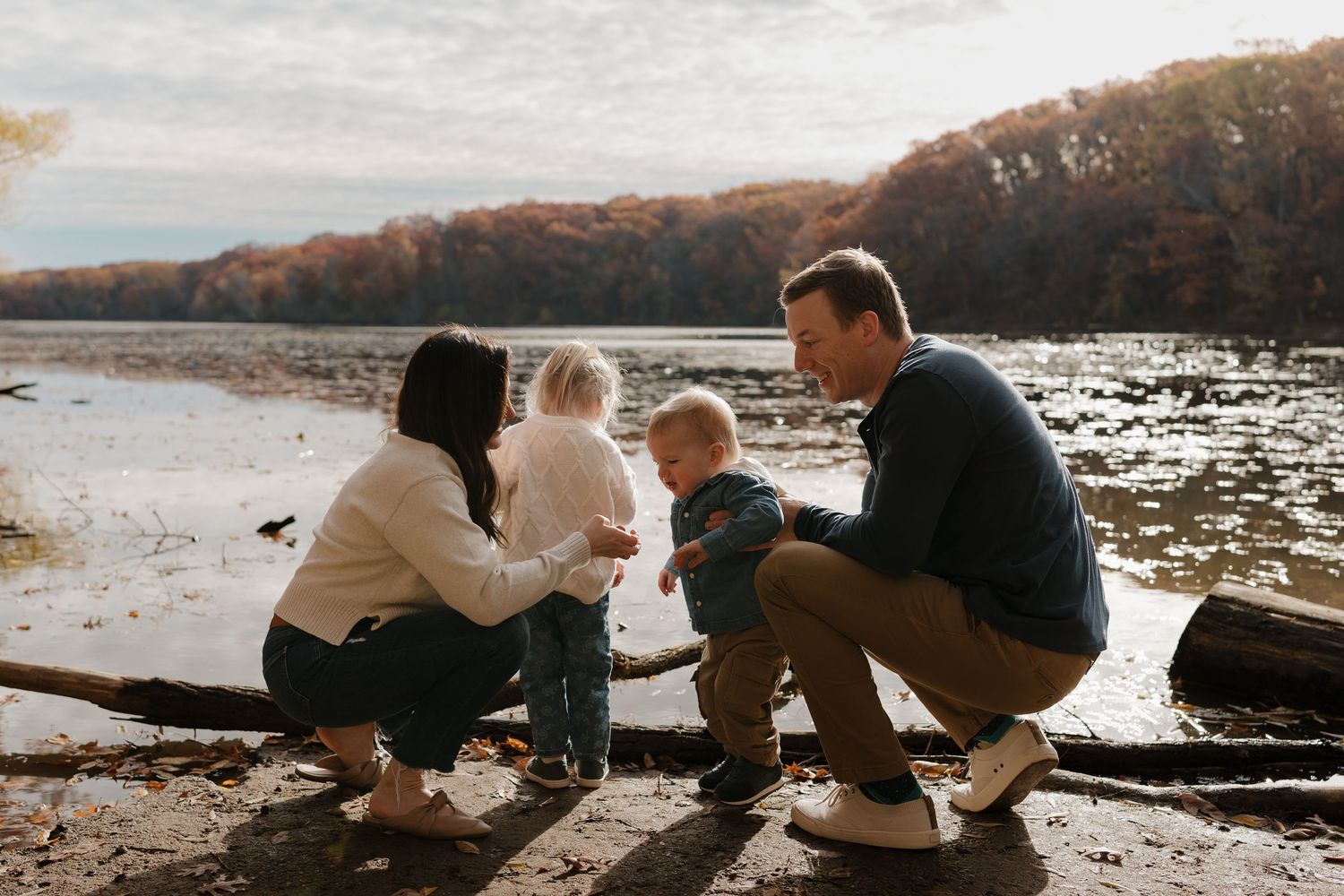 Family with two toddlers crouching by a lake shore during fall season.