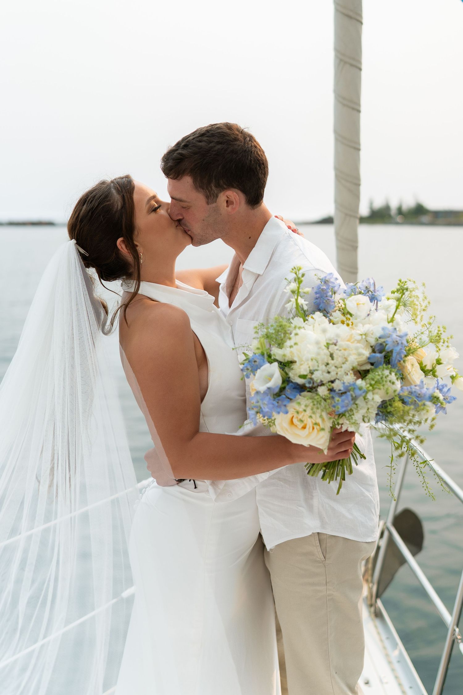 Bride and groom share a kiss on a sailboat, bride holding blue and white bouquet, wearing veil and white dress.