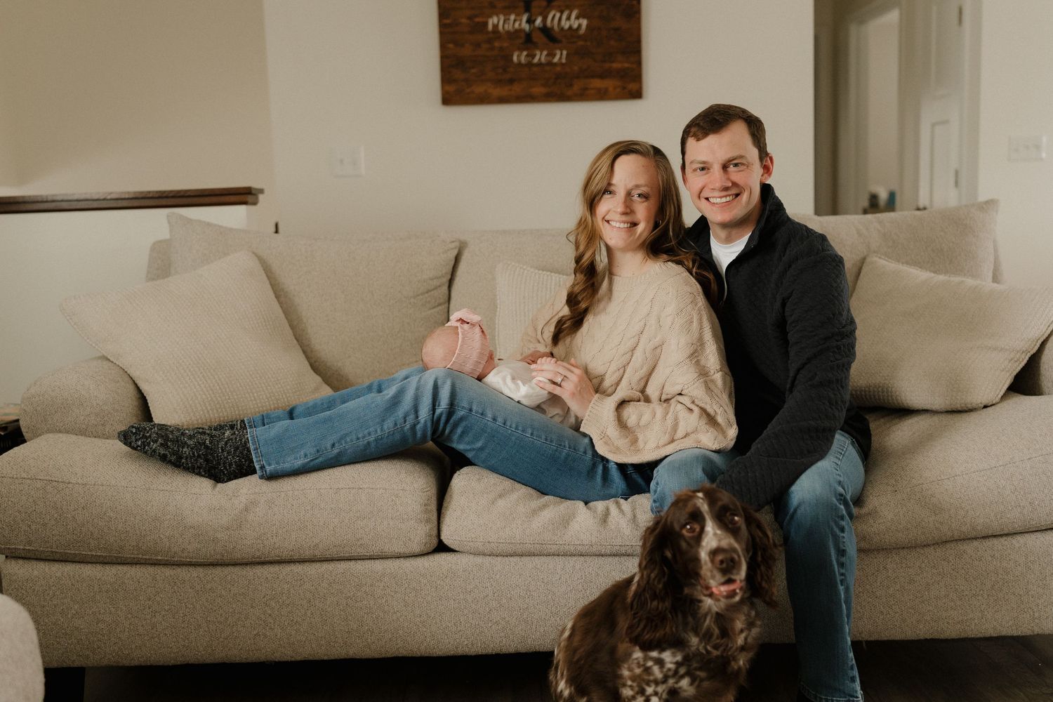 Couple sitting on beige couch with newborn baby and brown spaniel dog in cozy home setting.