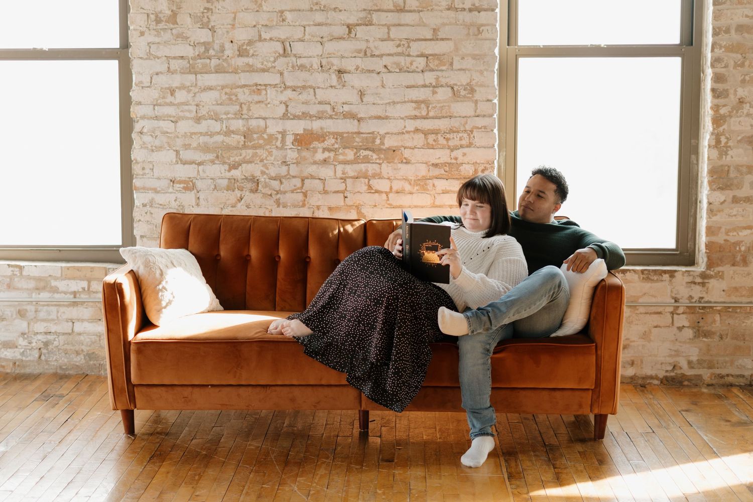 Couple relaxing on a velvet orange sofa in a bright loft apartment with exposed brick walls and hardwood floors.