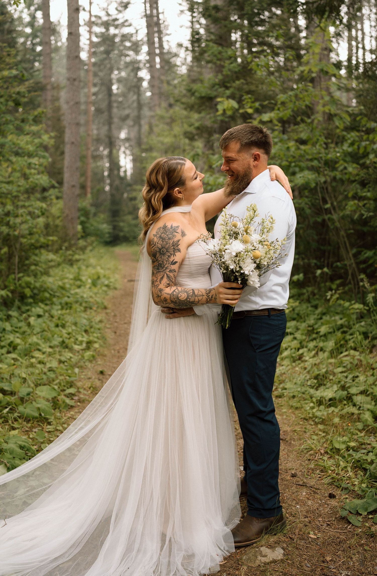 A bride in a white tulle gown with floral tattoos embraces her groom in navy pants on a forest trail.