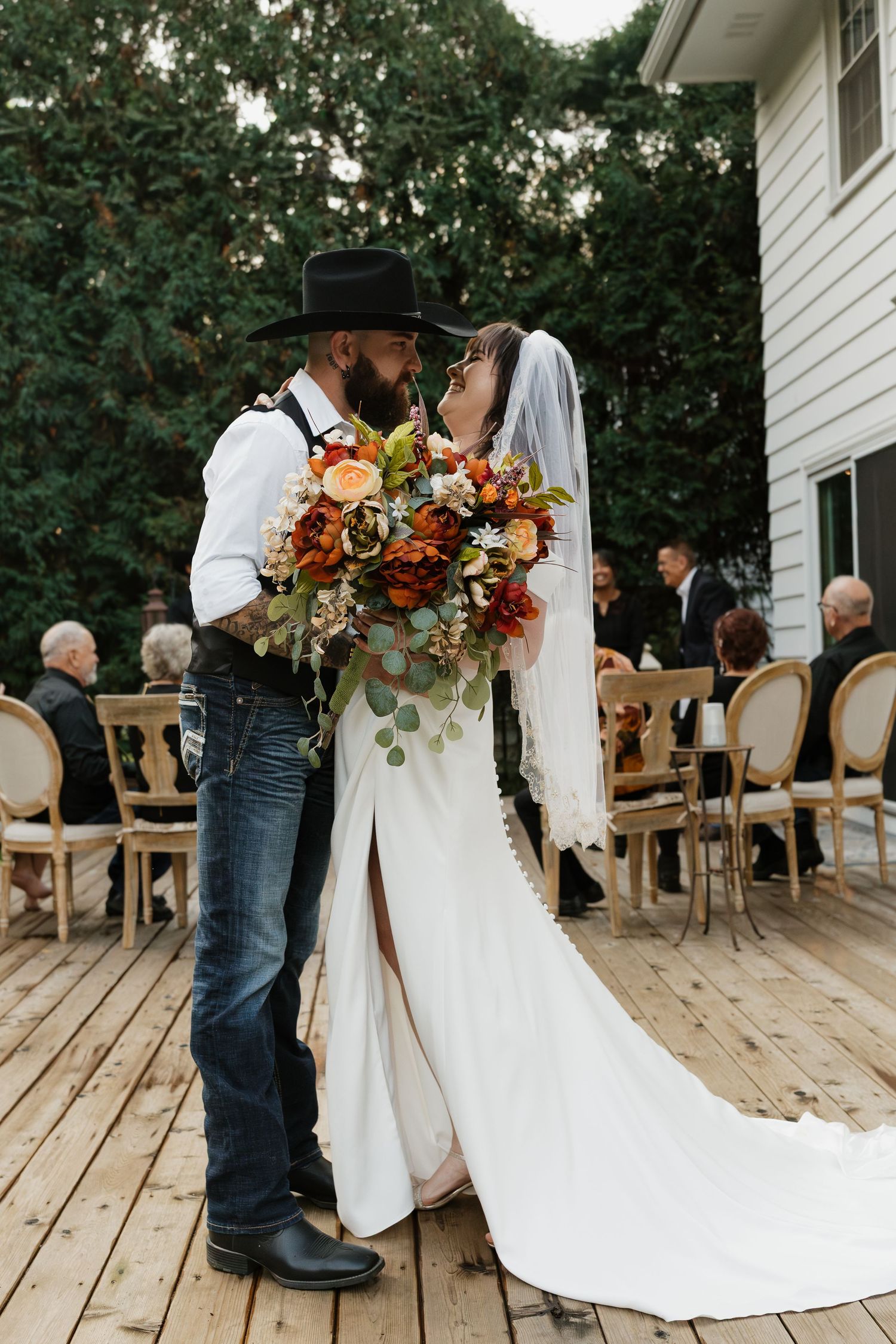 Newlyweds share a kiss on wooden deck, bride holding autumn bouquet, groom in black cowboy hat and jeans.