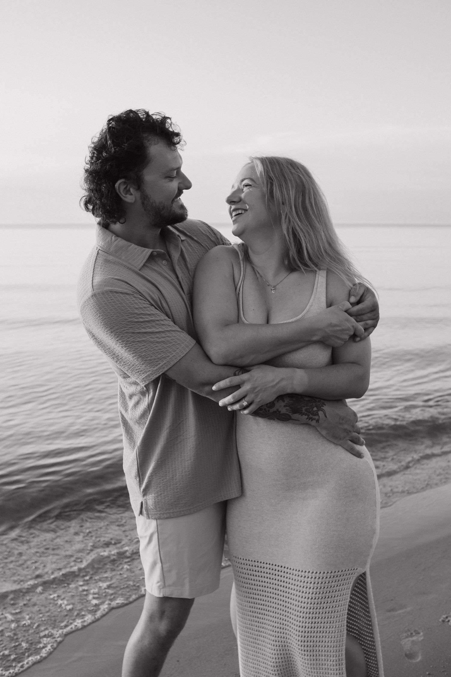 Black and white photo of a couple laughing and embracing on a beach with calm ocean waters behind them.