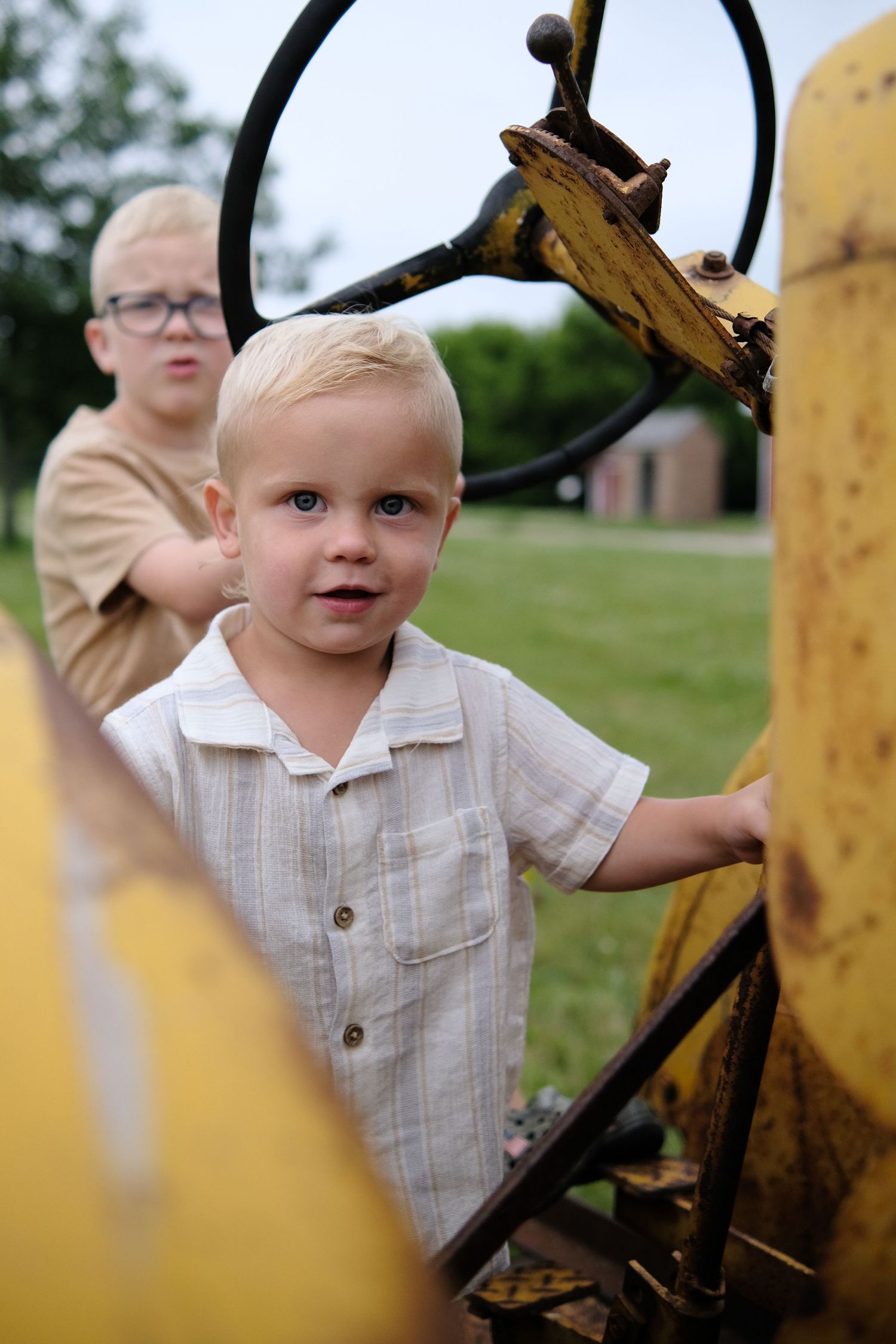 Two young blonde boys exploring vintage yellow tractor outdoors on green farm property.