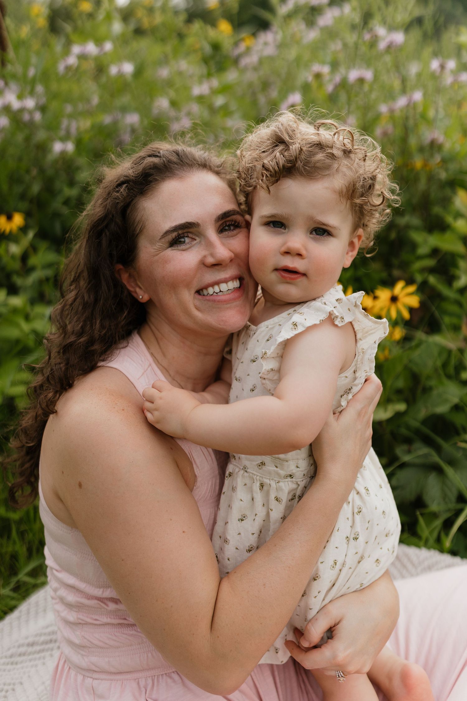 Smiling brunette mother hugs curly-haired toddler girl among yellow wildflowers outdoors.