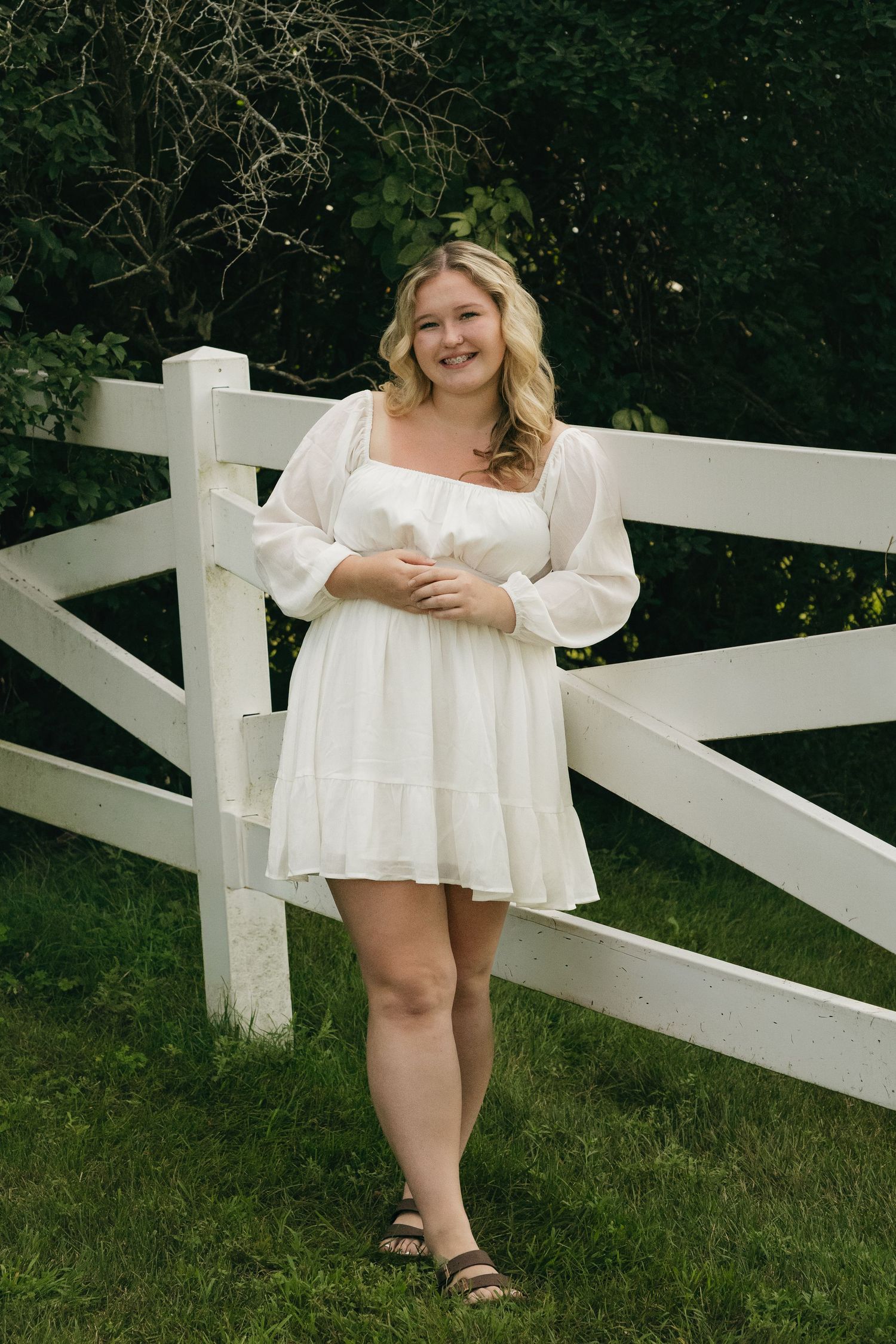 Smiling blonde woman in white dress stands by white wooden fence surrounded by greenery.