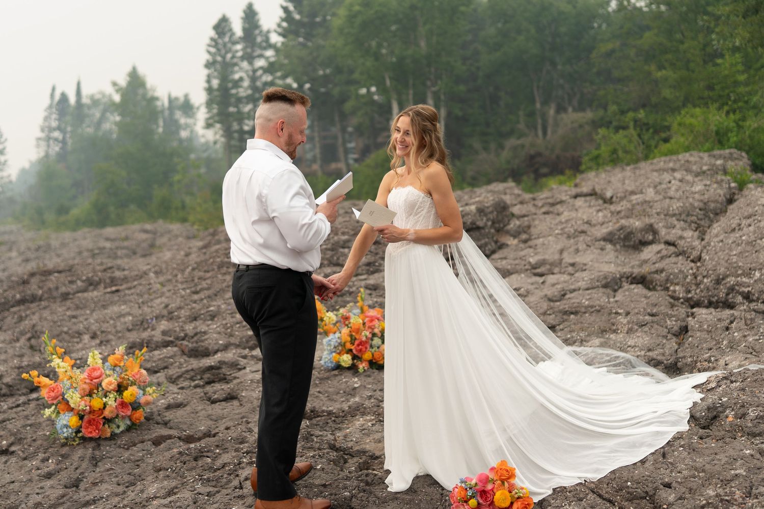 Couple exchanging wedding vows outdoors on rocky terrain with colorful floral arrangements and misty forest backdrop.