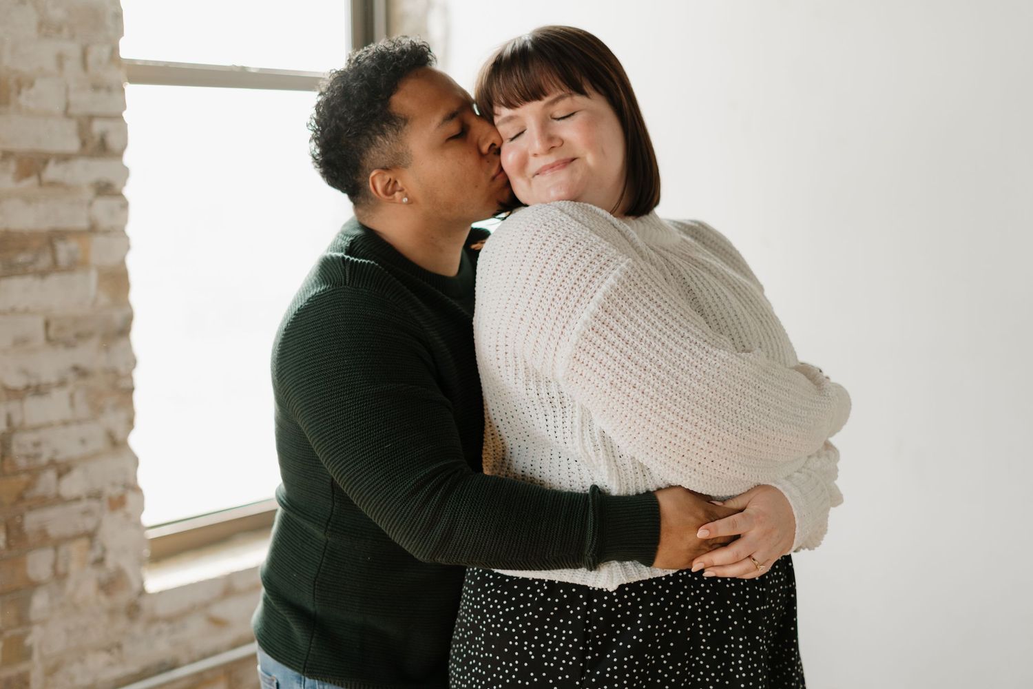 Two women sharing a warm embrace and kiss, one in a green top and one in a white sweater, standing near a bright window.