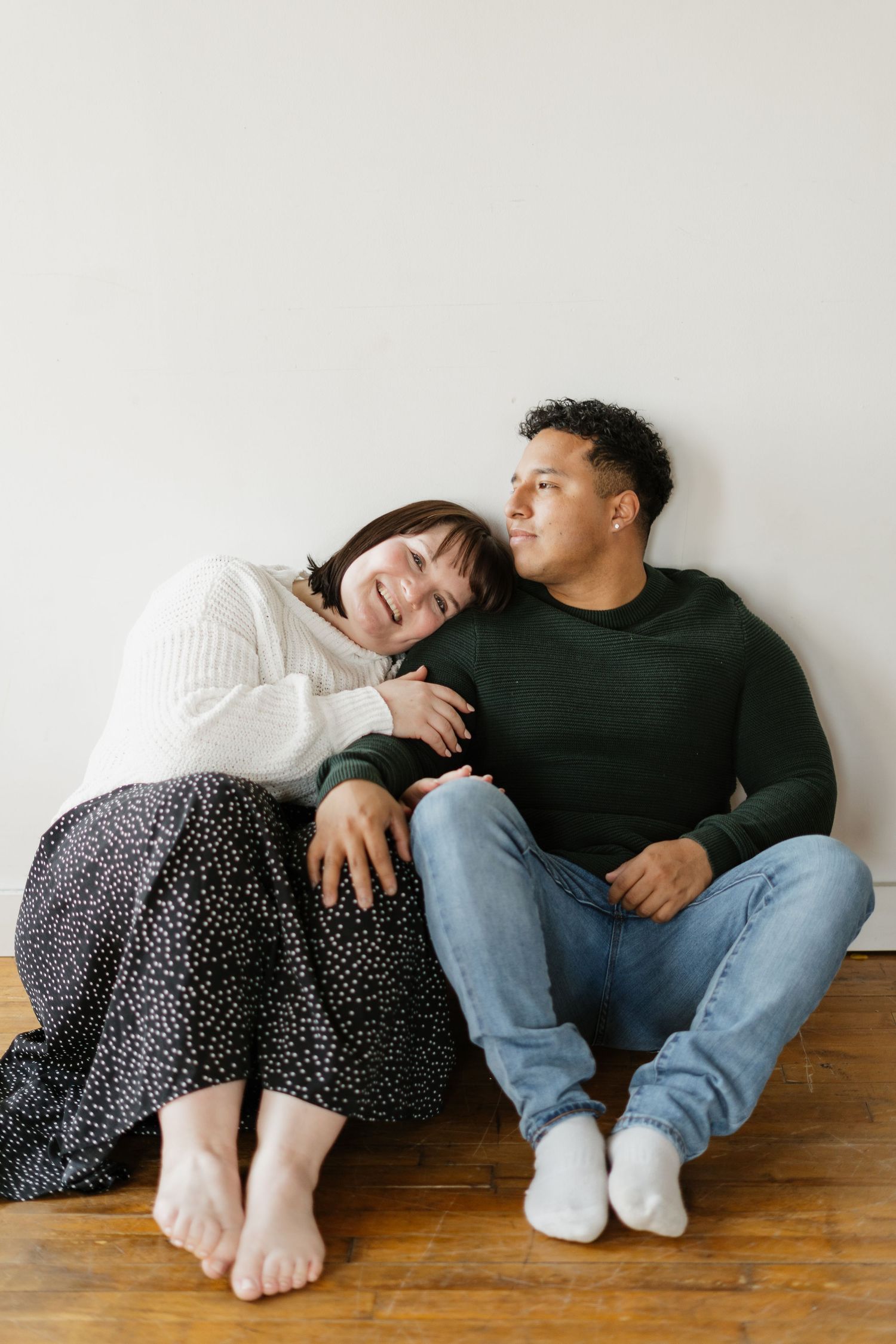 A smiling couple sits on a hardwood floor against a white wall, sharing a warm, affectionate moment together.