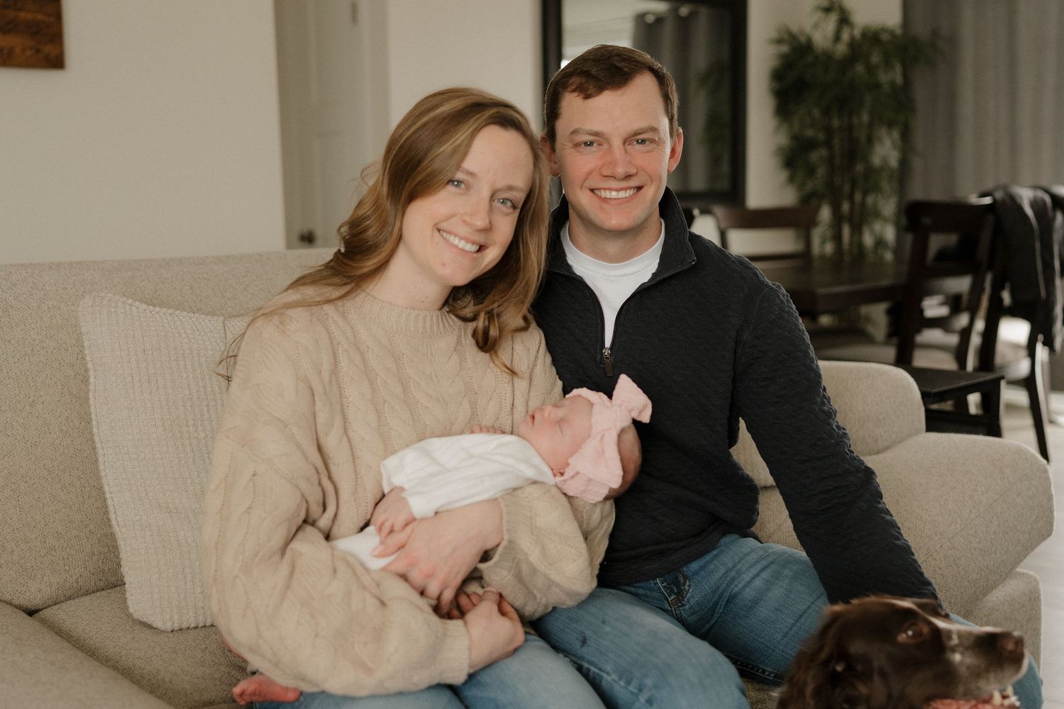 Young couple smiling on couch holding newborn baby with dog nearby in cozy home setting.