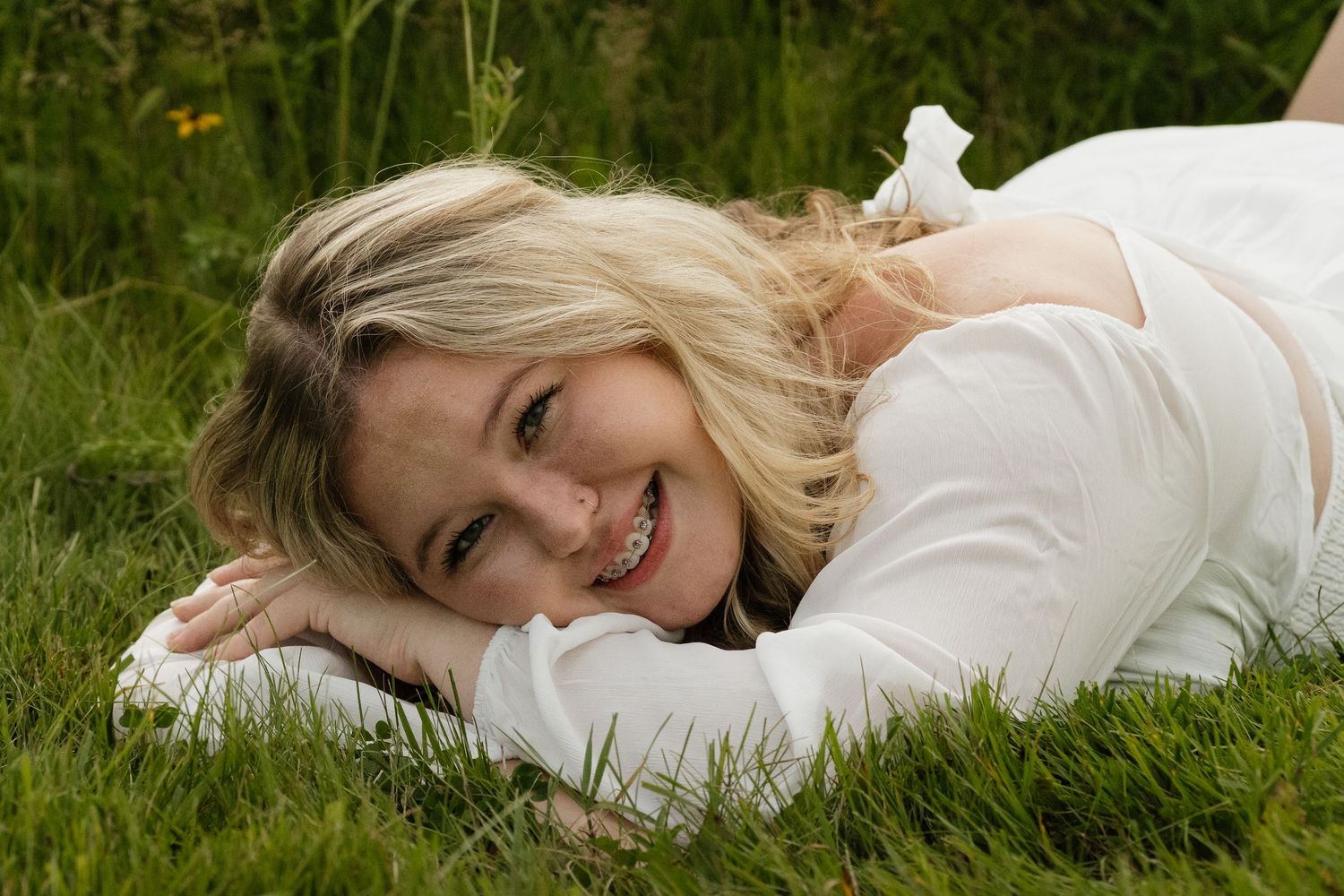 Smiling blonde girl in white dress lying on green grass looking up at the camera outdoors.