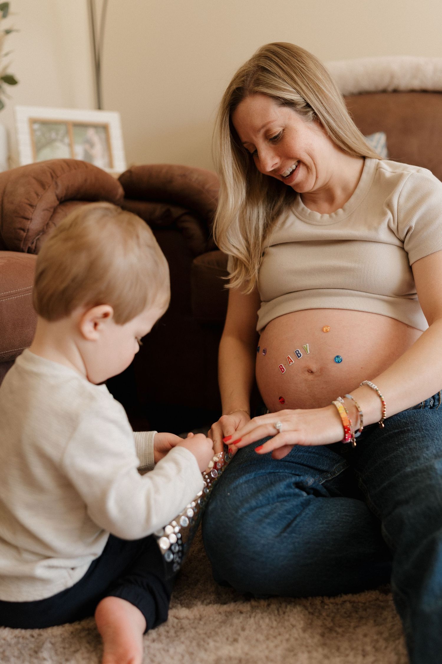 Pregnant woman smiling as toddler places stickers on her baby bump while sitting together on floor at home.
