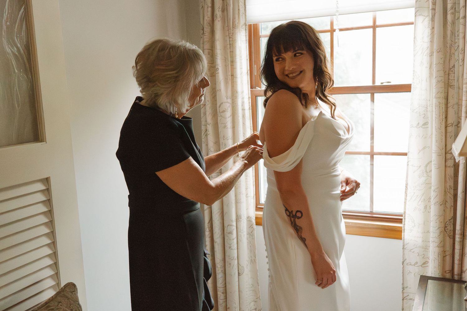 Older woman helping smiling brunette bride adjust her off-shoulder white wedding dress by window.