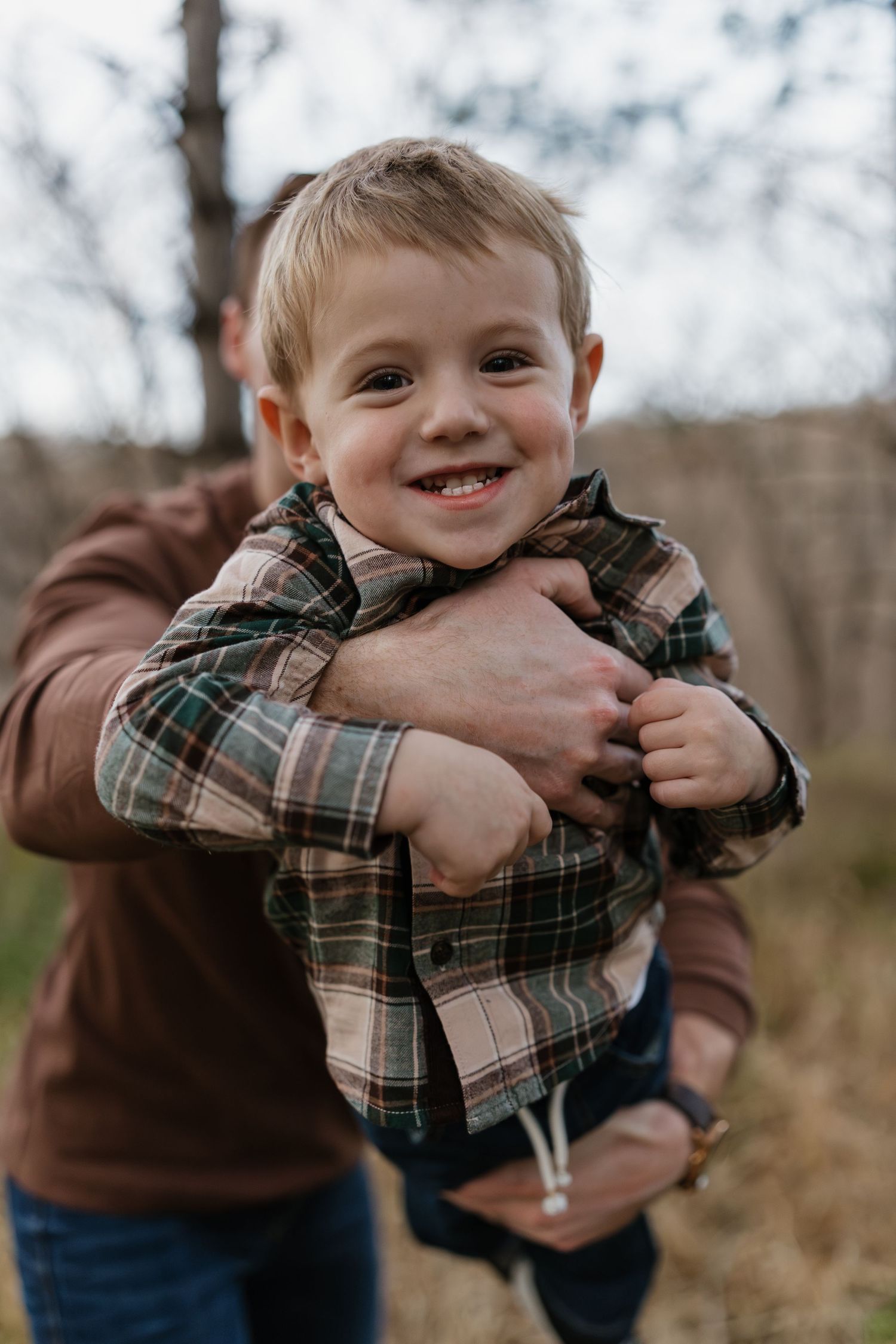 Happy toddler boy in plaid flannel shirt being held by father in brown sweater outdoors.
