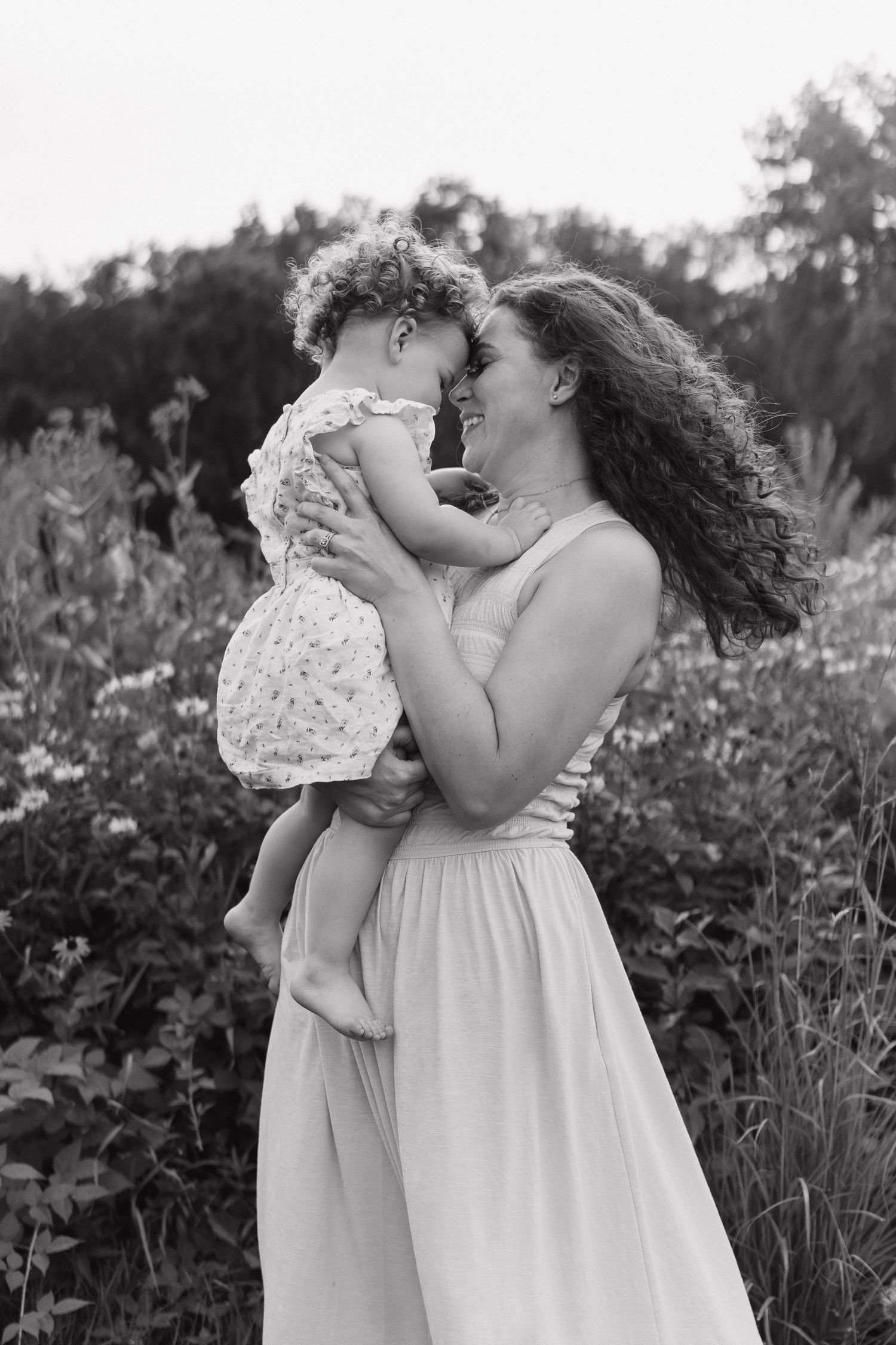 Black and white photo of a laughing mother holding her toddler nose-to-nose in a flower field.