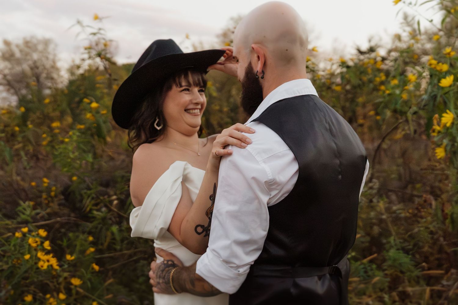 Bride in off-shoulder gown and black cowboy hat laughs with tattooed groom among yellow wildflowers.