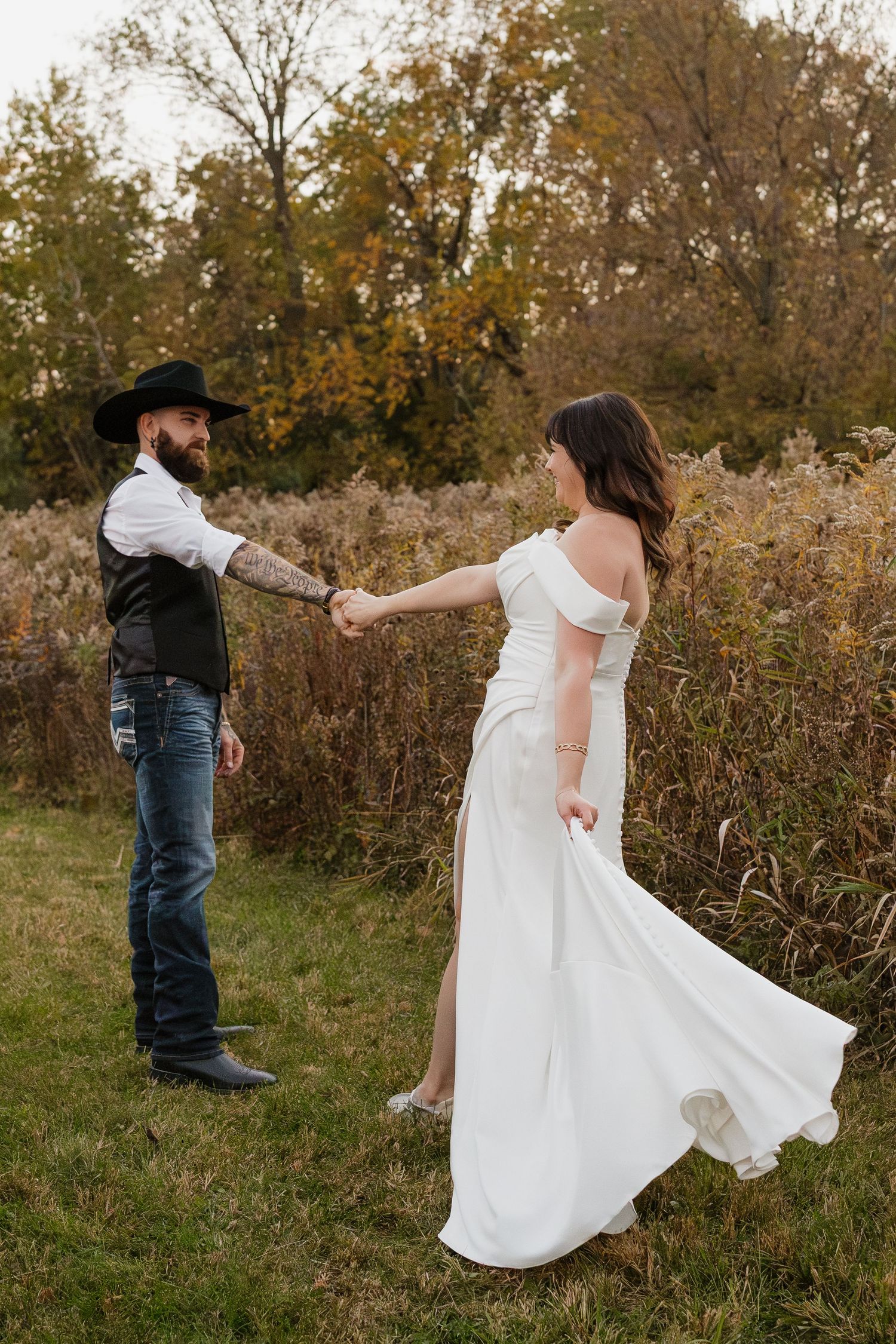 Bride in white off-shoulder gown holds hands with groom in cowboy hat and jeans outdoors in autumn.